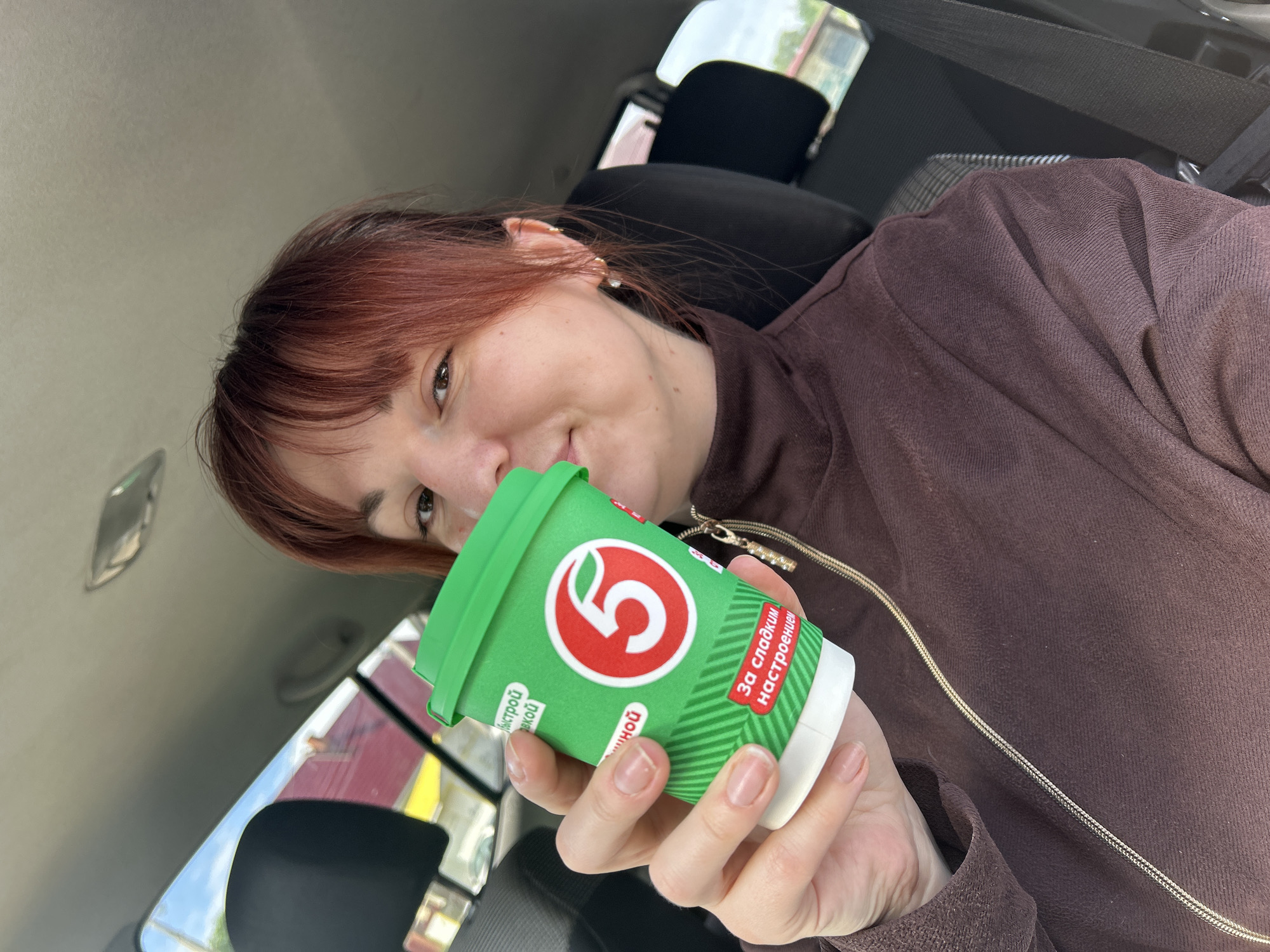 A woman with auburn hair is holding a green and white paper cup with a red number "5" logo, inside what appears to be a car. The text on the cup is in Russian. The woman is wearing a brown, long-sleeved top with a zipper and a charm. The background shows the car's interior, with a seat, seatbelt, and car roof visible. There's a glimpse of buildings and sky outside the car window. Based on the cup logo and text, the location is possibly in Фоки, Russia.