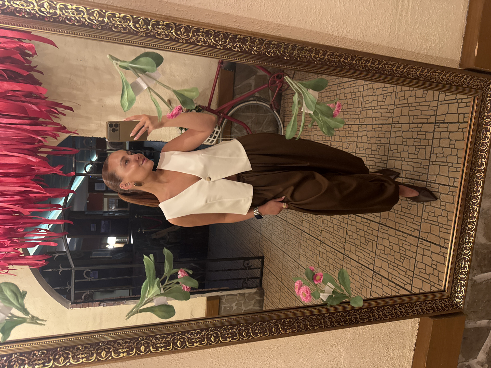 A full-length mirror selfie shows a woman standing indoors, positioned slightly to the left of the frame. She has reddish-brown hair pulled back into a ponytail and is holding an olive green or gold-colored smartphone with a triple camera array to take the picture. She wears a white, sleeveless, V-neck vest that fastens with a single light-colored button at the waist, and loose-fitting, dark brown, wide-leg pants. Her right foot, shod in a dark, pointed-toe shoe, is visible at the bottom of the mirror. She wears a silver-toned smartwatch on her left wrist and multiple thin rings on the fingers of her right hand. Dark, oblong earrings and a small helix piercing are visible on her left ear.
The mirror itself has a highly ornate, gold-colored frame featuring intricate scrollwork and floral patterns. Several artificial plant branches with green leaves and pink, daisy-like flowers are taped to the mirror's surface at various points.
In the reflection behind the woman, a light-colored, textured wall features an arched doorway with dark metal grates, leading into a dimly lit interior. To the right of the doorway, a red vintage-style bicycle with a white wicker basket on its handlebars is partially visible. The floor throughout the reflected space is covered in patterned tiles, forming a repeating geometric grid. Directly above the mirror, a dense cluster of red fabric strips or ribbons hangs from the ceiling. The visible walls outside the mirror frame are also light-colored and textured, with a wooden baseboard running along the bottom.