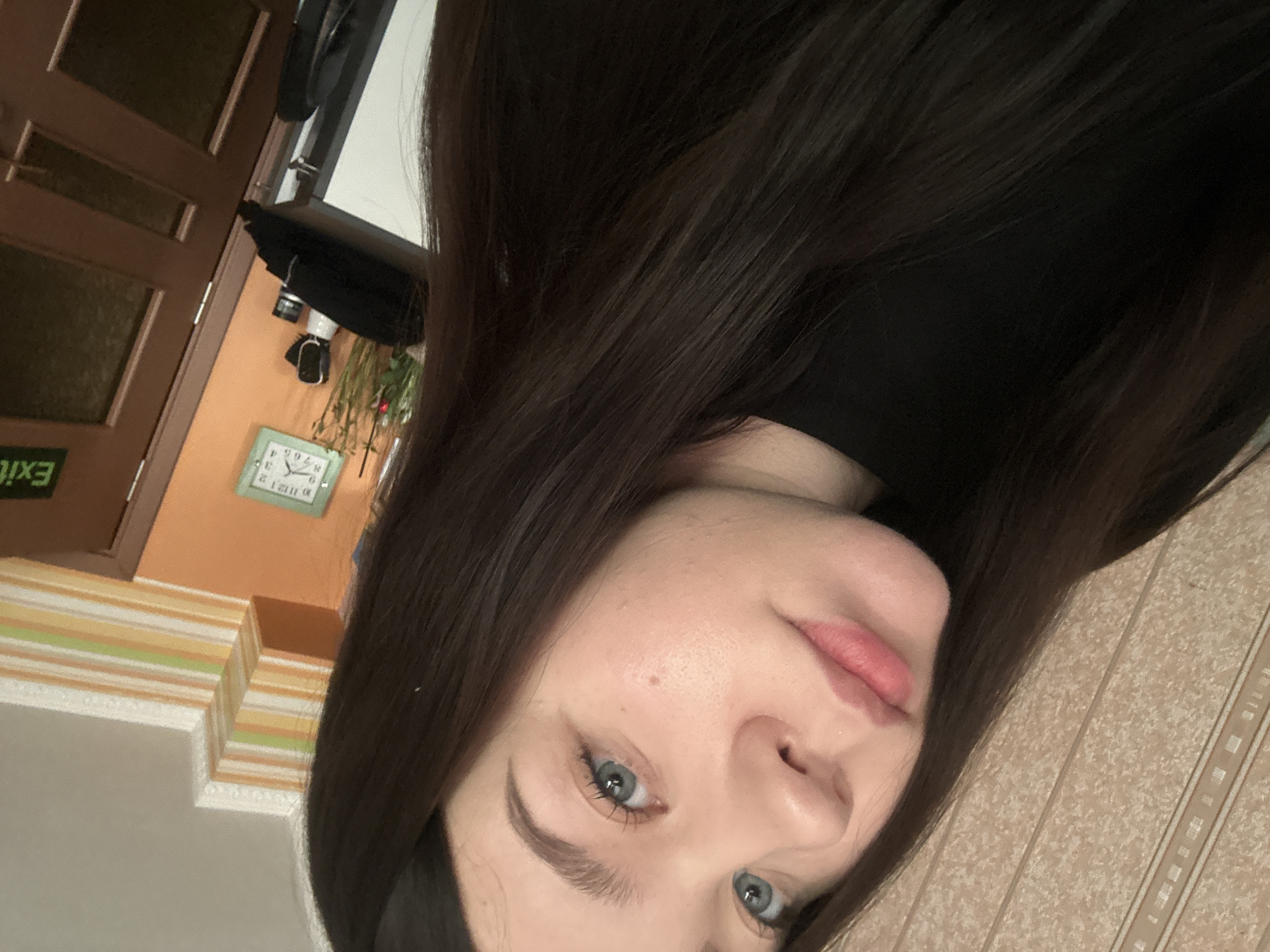 A close-up, tilted portrait photograph features a young woman with long, dark brown hair and pale skin. She has prominent blue eyes and natural, lightly pink lips. Her face is mostly clear, with a small mole visible on her cheek. She is wearing a black turtleneck sweater.
The background consists of a mixed indoor setting. To the left, there's a door with an "EXIT" sign (letters reversed, suggesting the photo is mirrored), and a striped wall design. Further back, there's a wall painted in a peach tone and a clock. To the right of the woman, there is a patterned beige wall.
There is not enough information to provide the specific location in the city of Varna, Bulgaria.