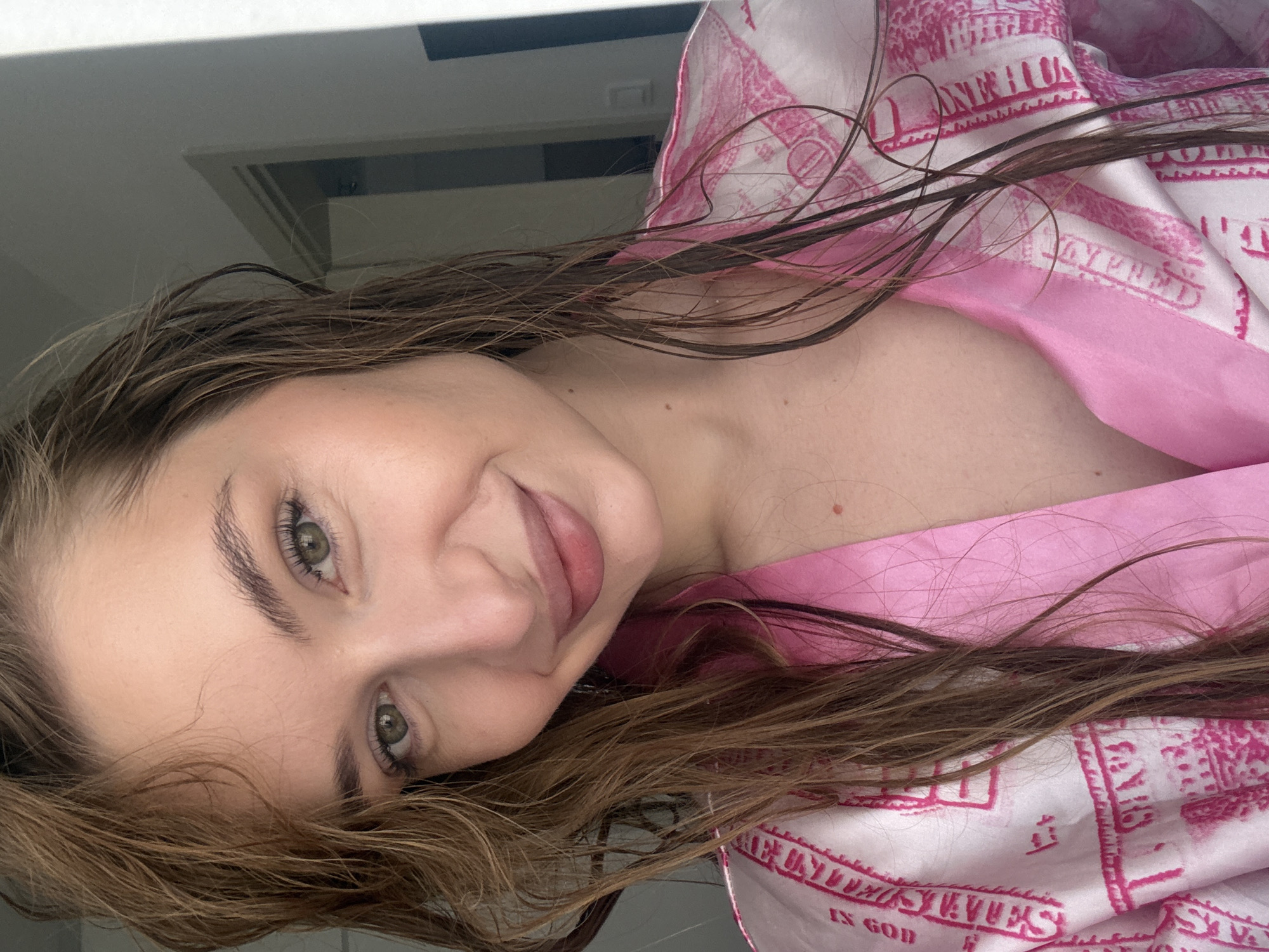 A young woman with shoulder-length brown hair and green eyes is depicted in an indoor setting. She is wearing a pink and white patterned robe and is looking at the camera with a slight smile. The lighting is soft and diffused. A small portion of a window or mirror is visible in the background. The location and context are not determinable from the image itself and do not suggest it is taken in Budva, Montenegro.