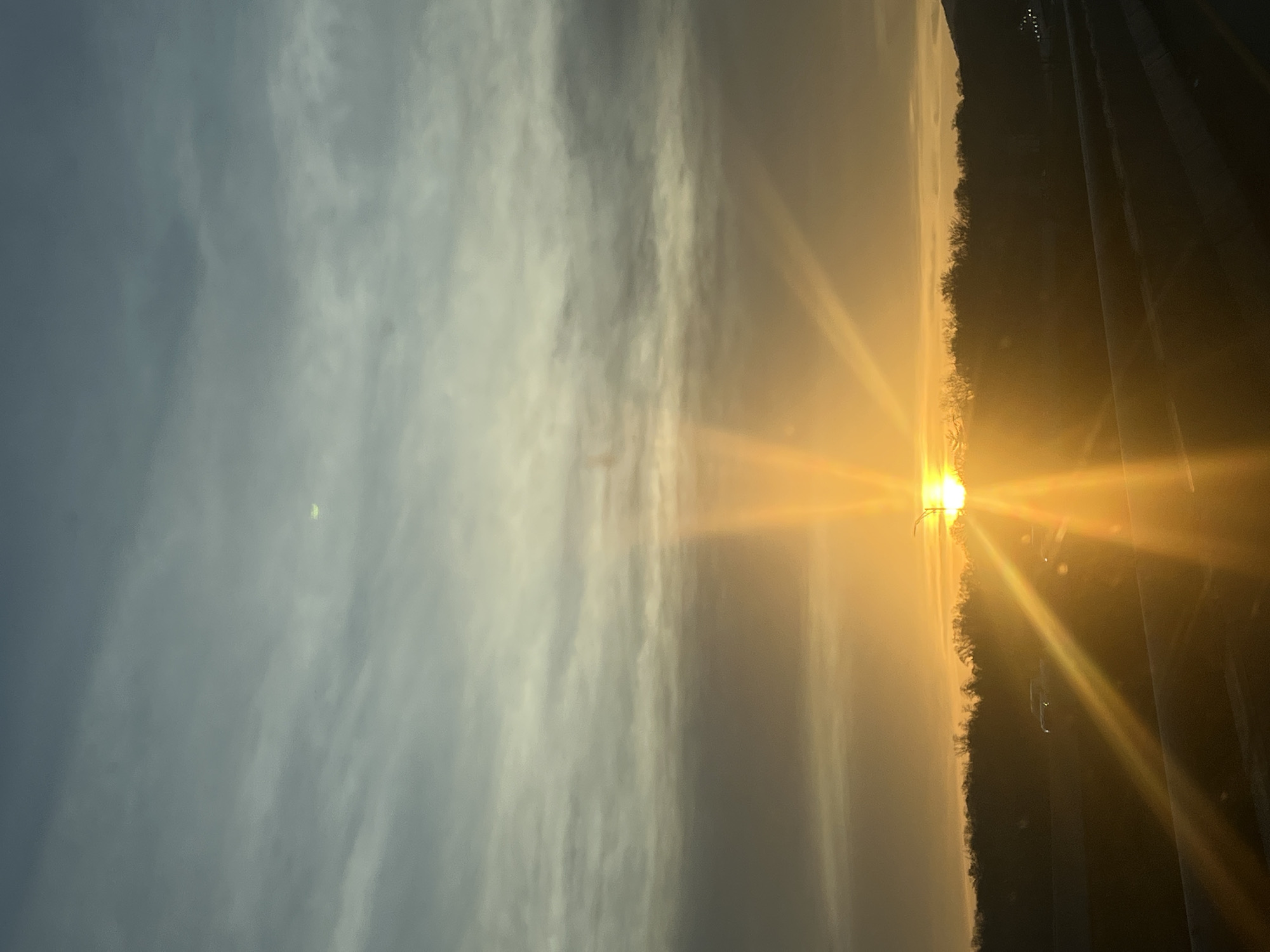 The image shows a sunrise in what appears to be a somewhat rural or suburban area. The sun is bright and low on the horizon, casting strong rays of light. Silhouetted trees are visible on one side, and a highway is seen on the right. The sky is partly cloudy with a mix of gray and lighter-colored clouds. It is not possible to pinpoint the specific location within the City of Newburgh or provide more detail about people or interactions, given the overall composition.
