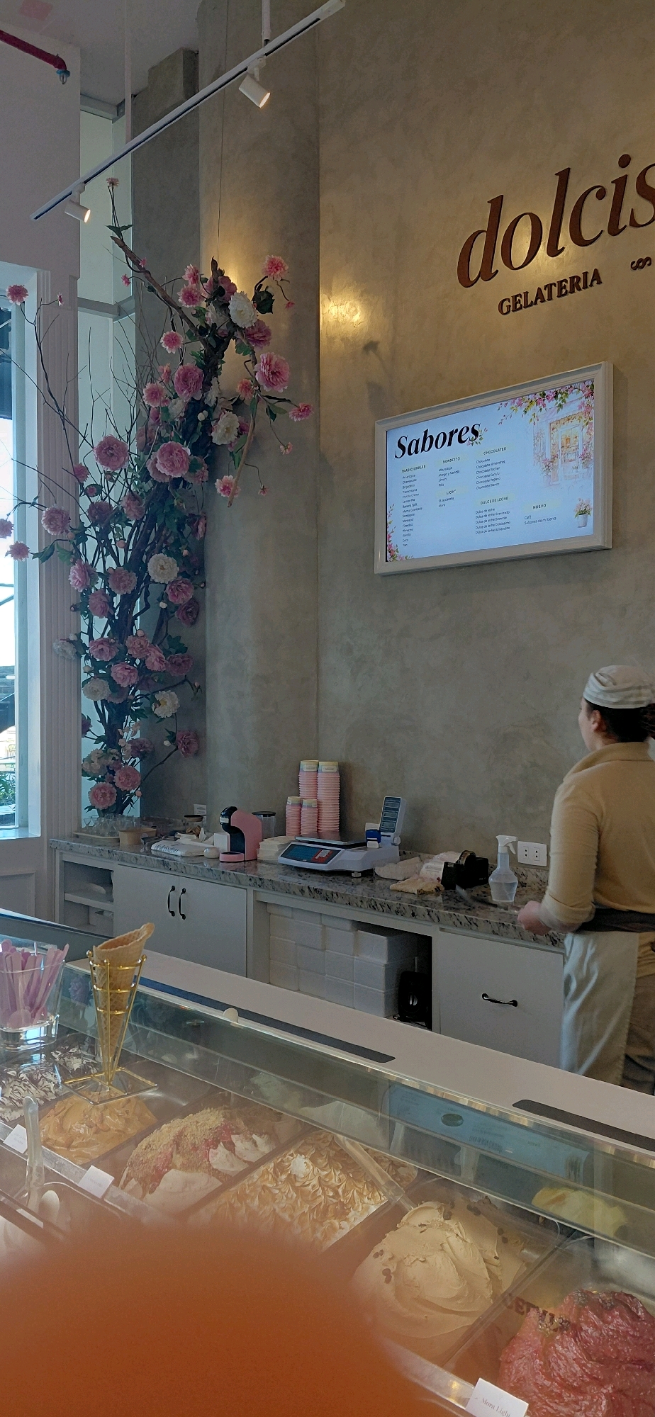 The image depicts the interior of a gelateria in Encarnación, Paraguay. The walls have a textured, grey finish, and overhead, a track lighting system illuminates the space. A large decorative floral arrangement, featuring pink and white flowers, is mounted on a branch structure on the left side of the image.
On the wall, a digital menu board displays "Sabores" (Flavors) at the top, with lists of ice cream flavors and categories such as "TRADICIONALES," "BOMBONSITO," "CHOCOLATES," and "DULCE DE LECHE." The gelateria's name, "dolcis GELATERIA," is prominently displayed in gold lettering above the menu.
A counter area is visible, equipped with a pink espresso machine, a digital scale, and stacks of pink cups. In the foreground, a display case showcases various flavors of gelato. A hand-held ice cream scoop and a golden cone holder are also present.
A woman, wearing a light-colored uniform and a cap, is partially visible in the right side of the frame, facing away from the camera and towards the counter. She appears to be an employee. The overall impression is of a clean, modern, and inviting establishment.