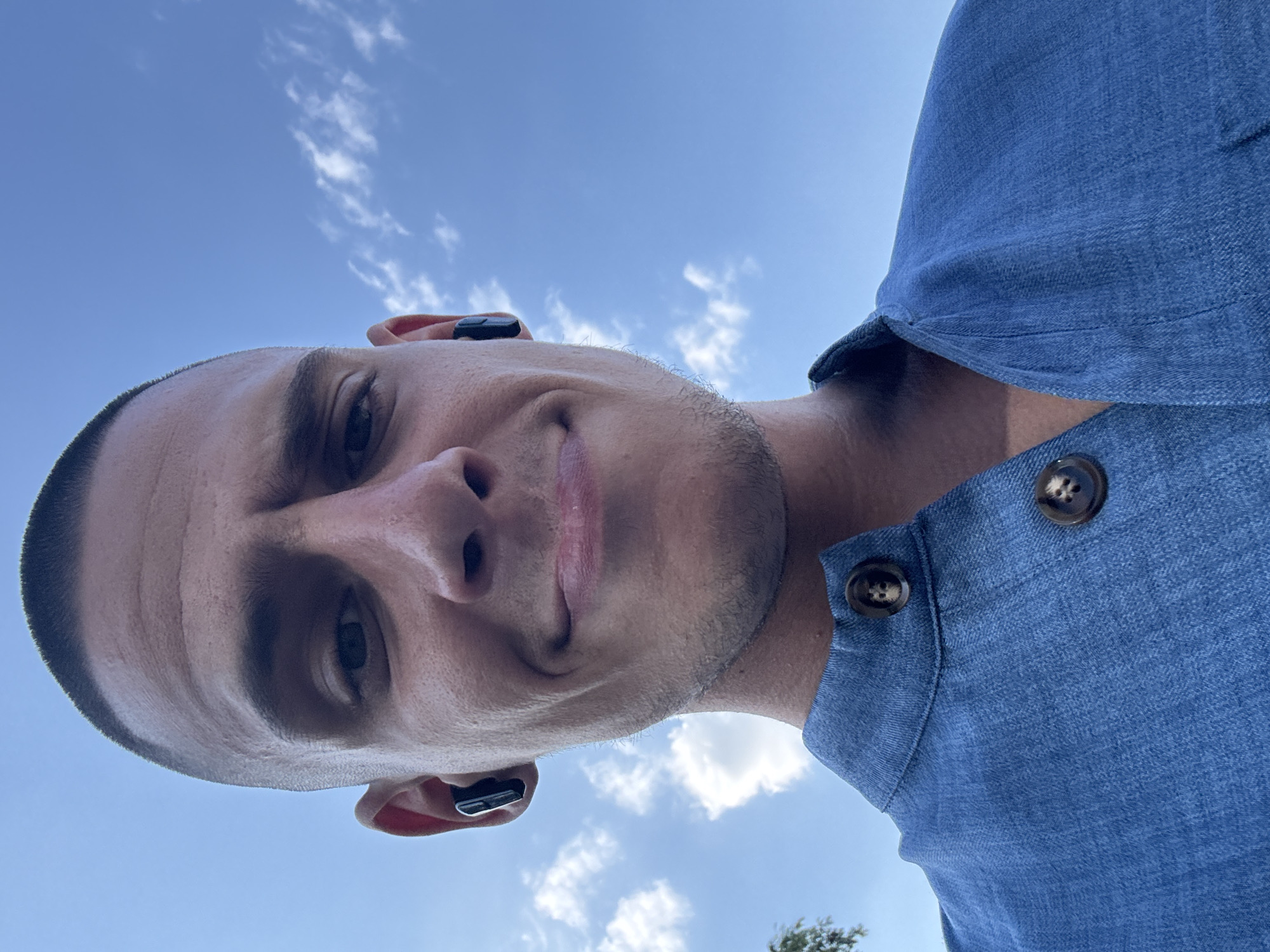 A close-up, low-angle, outdoor selfie shows a man with a buzzed haircut smiling faintly against a bright blue sky with scattered white clouds. He is wearing a blue collared shirt with two dark buttons visible on the placket. He has black wireless earbuds in both ears. The man appears to be in Sofia, Bulgaria, based on the provided context. The focus is on his face, with the sky dominating the background.