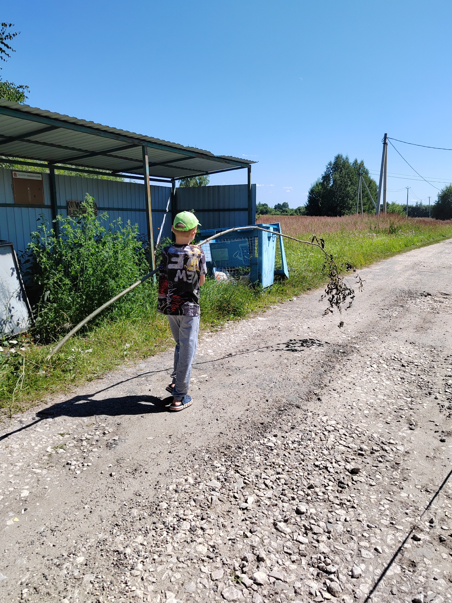 A young person wearing a green cap, a dark patterned shirt, and grey pants stands on a dirt road, holding a long stick. The stick is angled across their body, with dry leaves attached to one end. Behind the person, a blue dumpster sits near a covered structure with a metal roof. A metal fence encloses the area. The background features a field of dry grass, trees, and a clear blue sky. The road is covered in gravel and dirt, with shadows cast by the person and the stick. Power lines and a utility pole are visible in the distance.