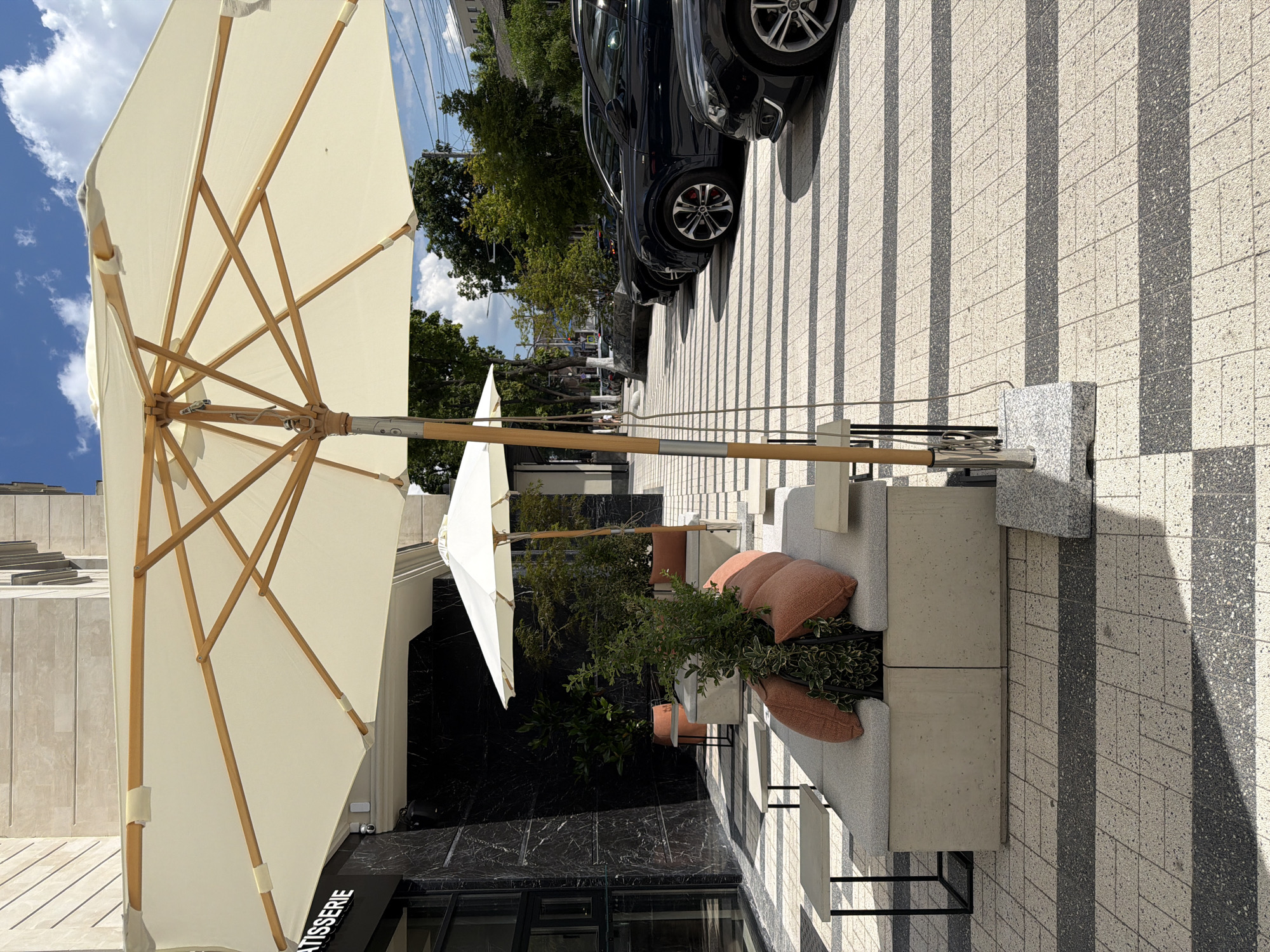 The image shows an outdoor seating area with umbrellas in front of a building in Chișinău, Moldova. A large white umbrella is prominent in the foreground, with its wooden frame visible. Beneath it, a modern concrete bench with orange cushions is set up. To the left, a building facade features dark marble-like cladding and the word "TISSERIE" is visible on a sign. The ground is paved with a pattern of light and dark rectangular tiles, with long shadows cast by the umbrellas and possibly other unseen objects. In the background, several dark-colored cars are parked in a row along the street. Green trees and a blue sky with white clouds complete the outdoor environment.