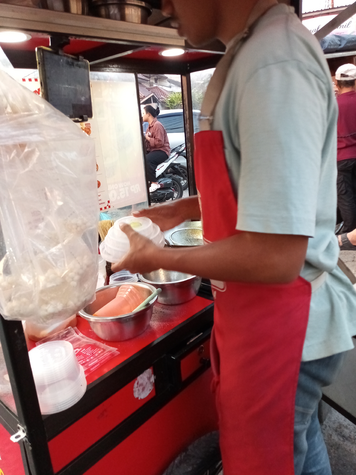 In Sariwangi, Indonesia, a person wearing a light blue t-shirt and a red apron stands behind a food cart. The cart has a red exterior with black accents. On the cart, there are several items including clear plastic bags filled with food, metal bowls, and stacks of white plastic containers. The person is holding a white plastic lid with a yellow object inside. In the background, a woman is sitting on a motorbike, and another person wearing a red shirt and white cap is visible. The scene appears to be outdoors, possibly on a street.