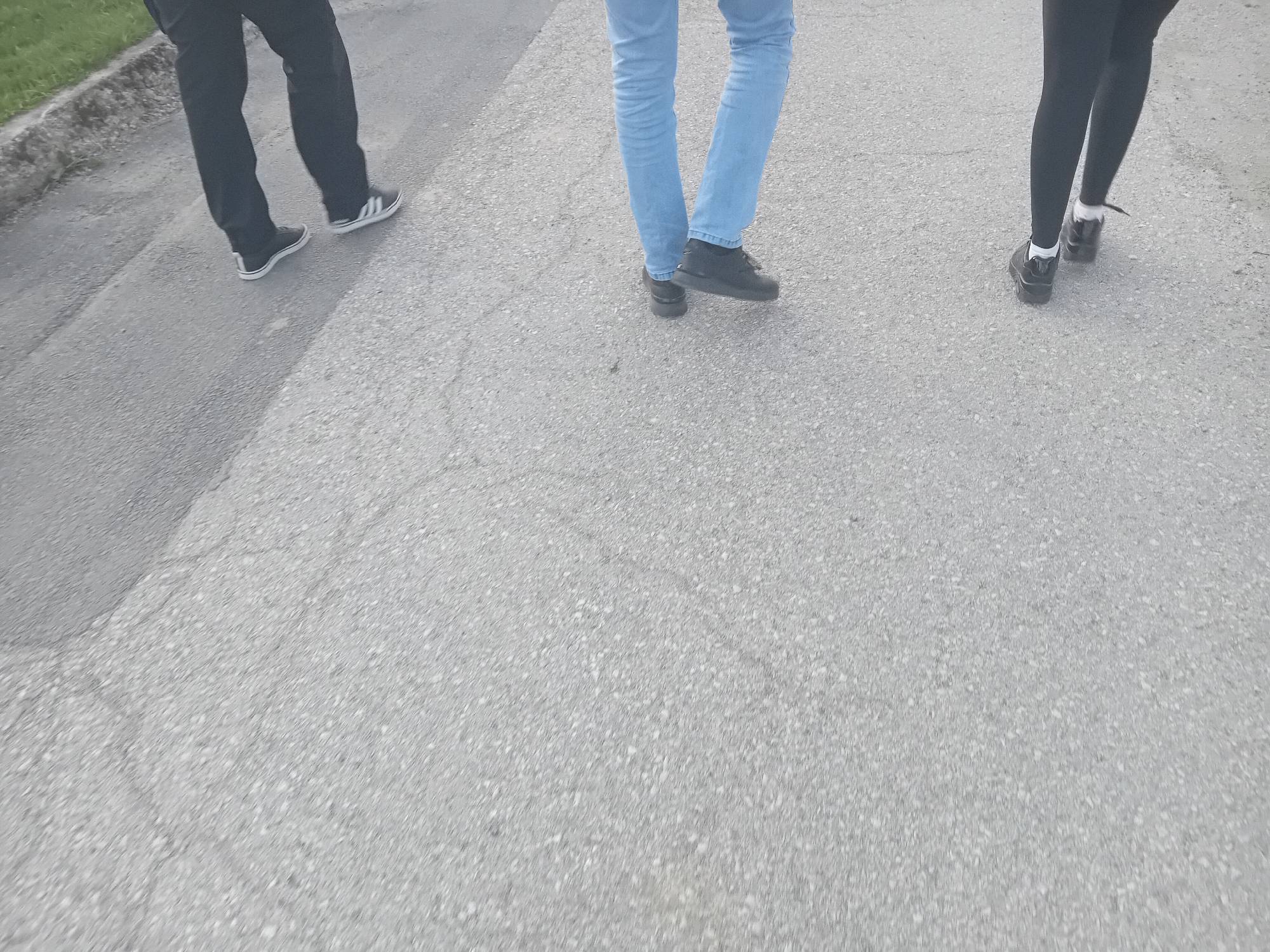 The image shows a low-angle, outdoor scene depicting the lower torsos and legs of three individuals walking on an asphalt surface. The individual on the left wears dark trousers and black-and-white athletic shoes. The person in the center wears blue jeans and dark shoes. On the right, an individual wears dark leggings and dark shoes with white socks. The background consists primarily of the asphalt surface and a blurred curb or edge in the upper left corner, potentially indicating a sidewalk or road. The location appears to be in an urban or suburban setting in the city of Unknown, Unknown.