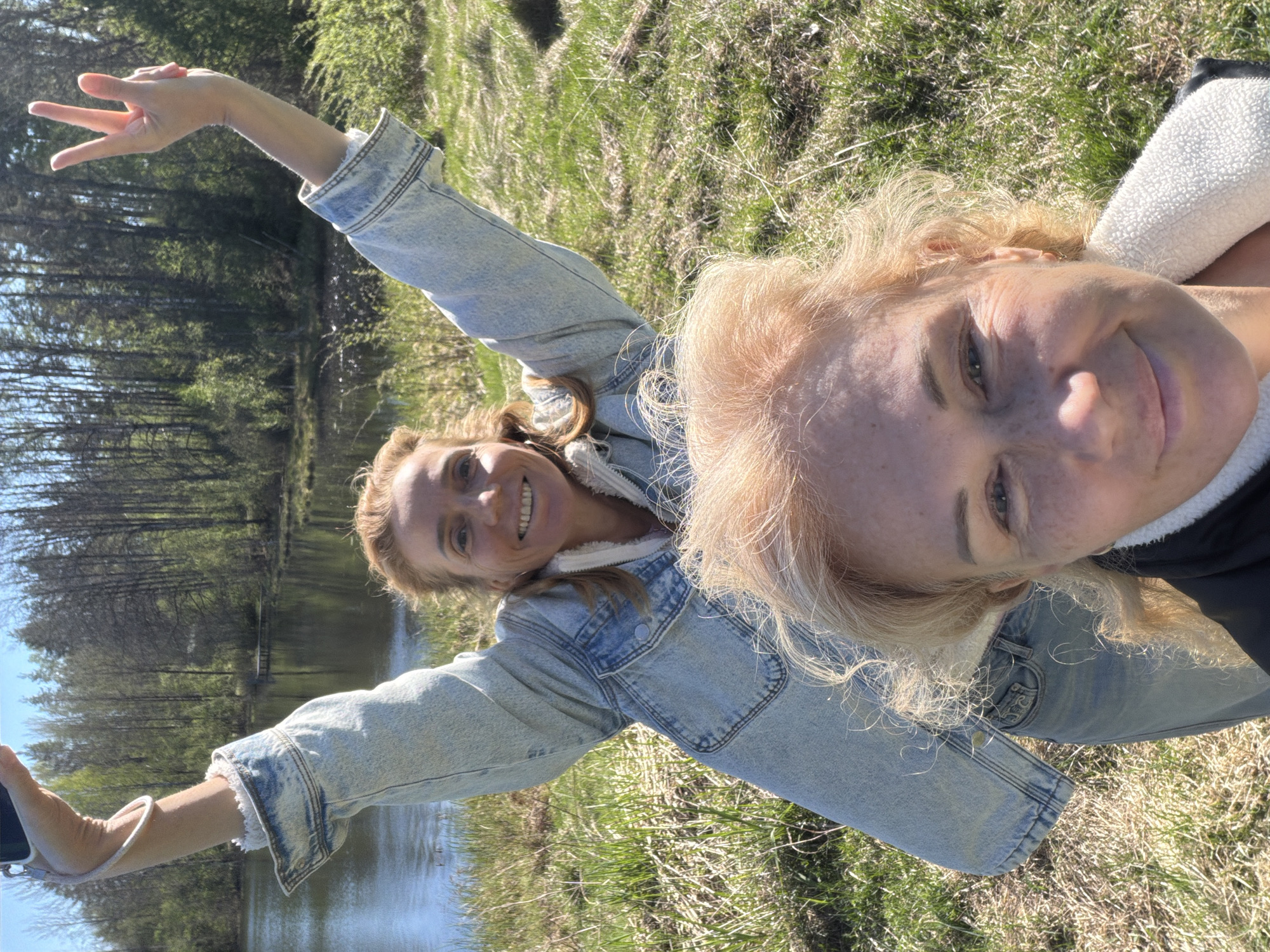 Two light-skinned women are posing for a selfie outdoors. One woman is blonde with fair skin, positioned closer to the camera with a slight smile. The other woman is also blonde and smiling, standing at a slight distance, wearing a light blue denim jacket with a white sherpa lining. Her left arm is extended upward in a V shape, and she is holding a cell phone in her right hand. They are standing near a body of water, possibly a lake or river, surrounded by a grassy area with trees visible in the background. The scene appears to be in a natural setting. It is daytime, and the lighting is bright. The location is likely in or around Vaskelovo, Russia, given the request.