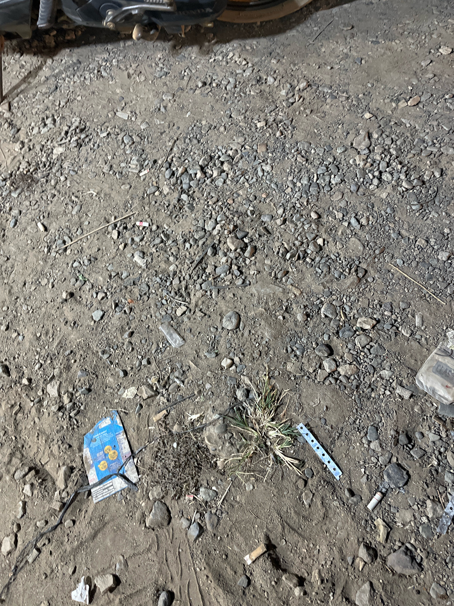 The image shows a patch of dirt and gravel ground with various objects scattered across it. In the upper portion of the image, a part of a motorcycle is visible, suggesting an outdoor environment. The ground is littered with small stones, dirt, and some debris. Among the debris, there are several cigarette butts, some appearing to be recently extinguished with smoke wisps. A crumpled blue and yellow paper object, possibly a wrapper or box, lies on the ground near the center-left. A small clump of grass is growing in the center of the frame. To the right of the grass, a thin, perforated strip of blue plastic is visible, with circular holes. A clear plastic bottle fragment is also present in the lower right quadrant. The lighting suggests it is either dusk or nighttime, with artificial light casting shadows. The scene appears to be an unkempt outdoor area in Kemuning, Indonesia.