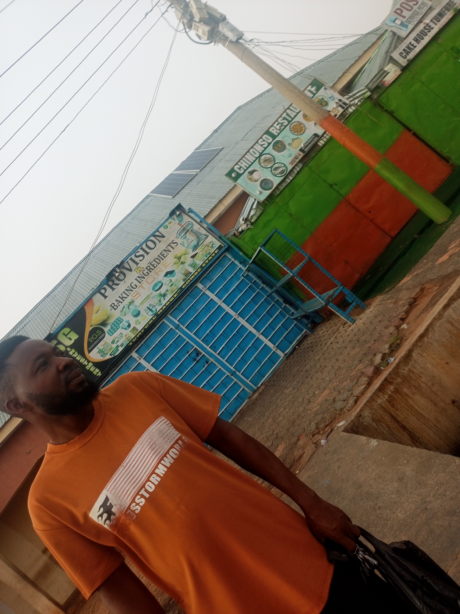 A man stands in front of a business complex in Tumfure, Nigeria. He is wearing an orange t-shirt with the inscription "SSTORMWOREA" and carrying a black plastic bag. Behind him, there are several businesses, including "PROVISION BAKING INGREDIENTS STORE," "CHINONSO RESTAURANT," and "POS CAKE HOUSE TUMFURE." The buildings are painted in a variety of colors, including blue, green, and red. A utility pole is visible in the background with solar panels on the roof.