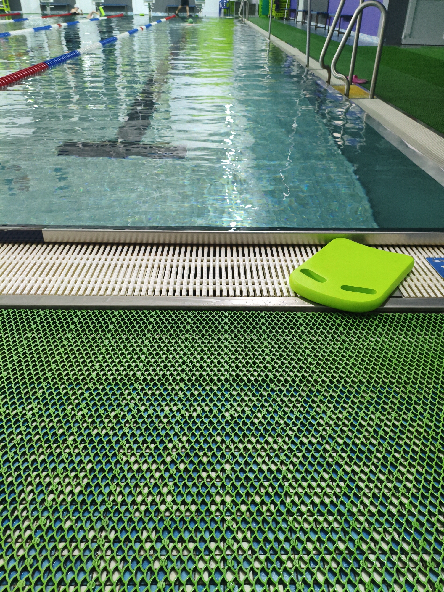 The image shows a brightly lit indoor swimming pool with clear blue water and lane markers. A vibrant green kickboard rests on a white slatted edge by the pool. In the foreground, a textured green mat with a fish-scale pattern covers the floor. The environment appears clean and well-maintained, suggesting a public or private aquatic facility.