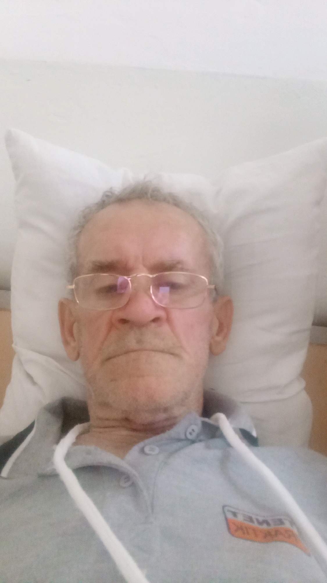 A man with graying hair and glasses is lying on a pillow, with his head slightly tilted. He is wearing a light gray polo shirt with white drawstrings. On the left side of his shirt, there is a logo that appears to be a white rectangle with orange lettering. The background is a plain white wall or surface. The lighting is bright and even, suggesting an indoor setting, possibly a bedroom or hospital room in Tiachiv, Ukraine.