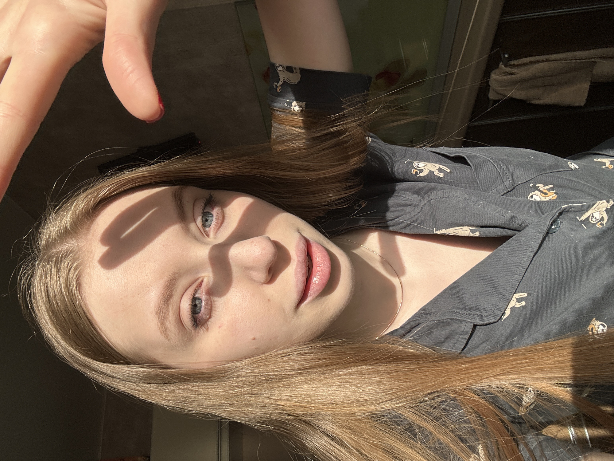 A young woman with light blonde hair and fair skin is featured in a close-up, slightly angled shot. She is wearing a dark button-up shirt with a light-colored cartoon pattern. The lighting is bright and sunny, casting shadows on her face from a hand held up above, suggesting an indoor environment. She appears to be in a bathroom. The subject is smiling at the camera. Based on the picture, there is no indication that the photo was taken in the city of Chaikovsky, Russia.