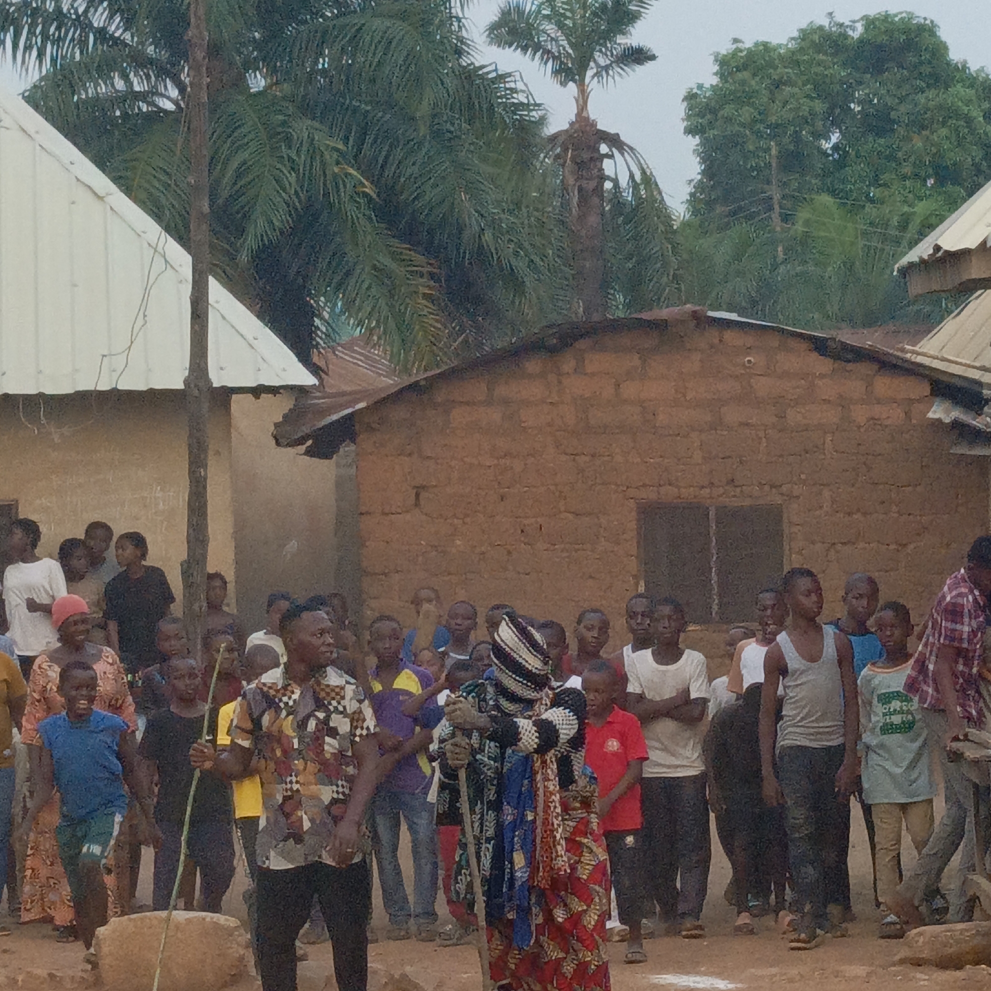 A medium shot depicts a gathering in what appears to be Akwanga, Nigeria. A crowd of people, mostly children and young adults, surround a central figure dressed in a traditional costume with black and white headgear, and a patterned cloak over a red skirt. The person holds a wooden staff. To the left stands a man wearing a patterned shirt and black pants, also holding a wooden stick. The setting includes traditional houses made of mud bricks with corrugated metal roofs. A palm tree is visible in the background. The environment suggests a rural or semi-rural community.