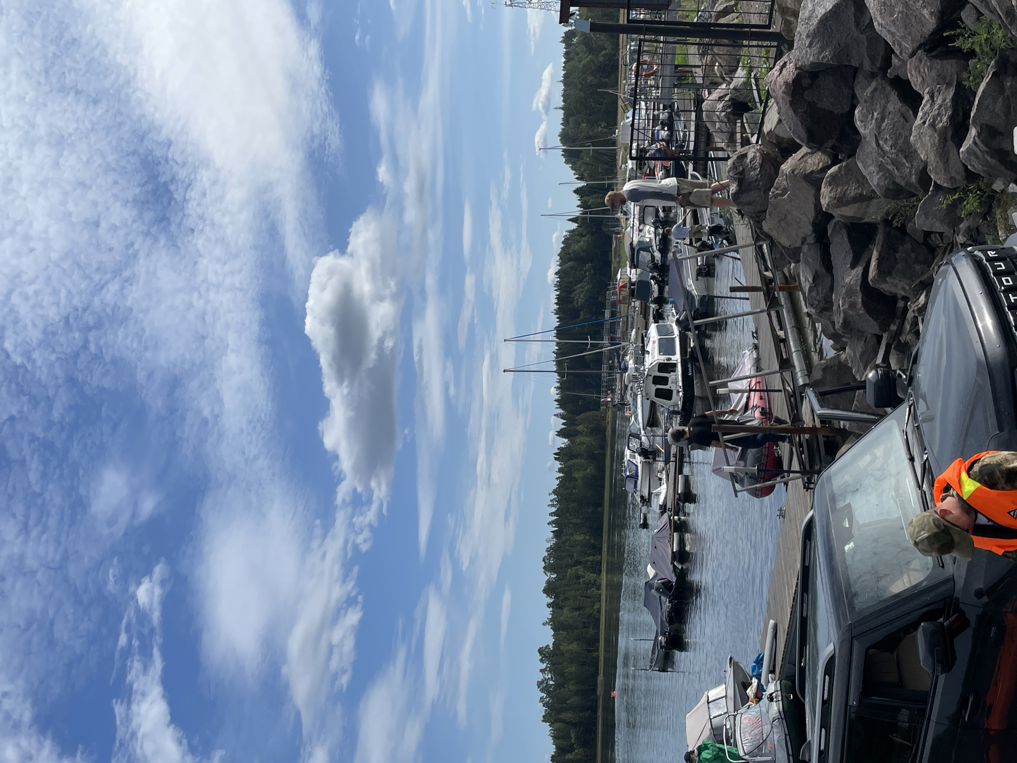 The image depicts a marina in Berozovo, Russia, under a bright blue sky with scattered white clouds. Several boats, ranging from small motorboats to sailboats, are docked in the water. The shoreline is lined with a dense forest of green trees. On the dock, several people are visible. One man, wearing a grey and white long-sleeved shirt and beige shorts, stands on a pile of rocks, holding a bag. Another man in a black shirt and blue jeans walks along the dock. In the foreground, a black SUV is parked, with a person wearing an orange safety vest and a camouflage hat visible in the driver's side. The scene suggests a recreational boating or fishing activity.