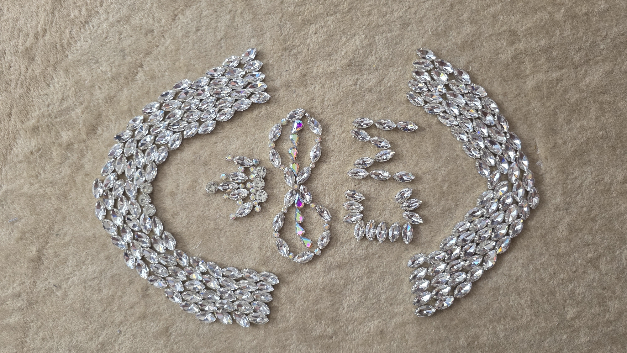 The image displays an intricate arrangement of numerous clear, faceted, marquise-shaped rhinestones and crystals on a light brown, soft, textured fabric surface, possibly faux fur or velvet. Two large, curved, wing-like patterns, formed by multiple rows of clear rhinestones, frame the central elements.
In the center, three distinct symbols or characters are meticulously formed by the crystals. From left to right, these appear to be:
1. A stylized Chinese character resembling '发' (fā), meaning "to prosper" or "to get rich," primarily formed with clear crystals.
2. An elongated infinity symbol or a stylized number '8,' crafted from a mix of clear and iridescent (AB finish) crystals, which show a subtle rainbow shimmer.
3. Another stylized Chinese character resembling '福' (fú), meaning "fortune," "blessing," or "happiness," composed entirely of clear crystals.
The combination of these symbols suggests a theme of enduring prosperity, wealth, and good fortune, potentially referencing the auspicious significance of the number eight (which sounds like "fa" 發, similar to "fā") in some cultures. The arrangement highlights the sparkle and facets of the individual stones against the soft background.