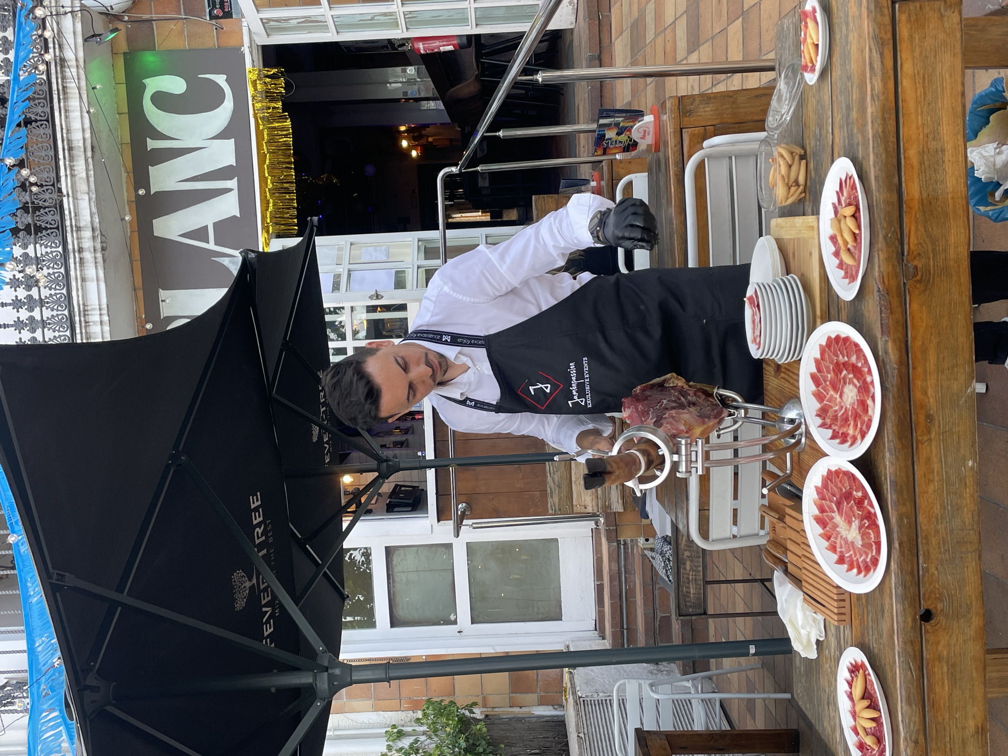 A man, wearing a black apron with the logo "Jamónpassion EXCLUSIVE EVENTS", is slicing a large cut of ham with a knife and fork. He is standing behind a wooden table laden with plates of thinly sliced ham and breadsticks. The scene appears to be outdoors, possibly at a restaurant or event. A large black umbrella, with the "FEVER TREE MIX WITH THE BEST" logo, provides shade. In the background, a sign reads "BLANC" in large white letters. The floor is paved with reddish-brown tiles. The overall setting suggests a food-related event or establishment in Arenys de Mar, Spain.