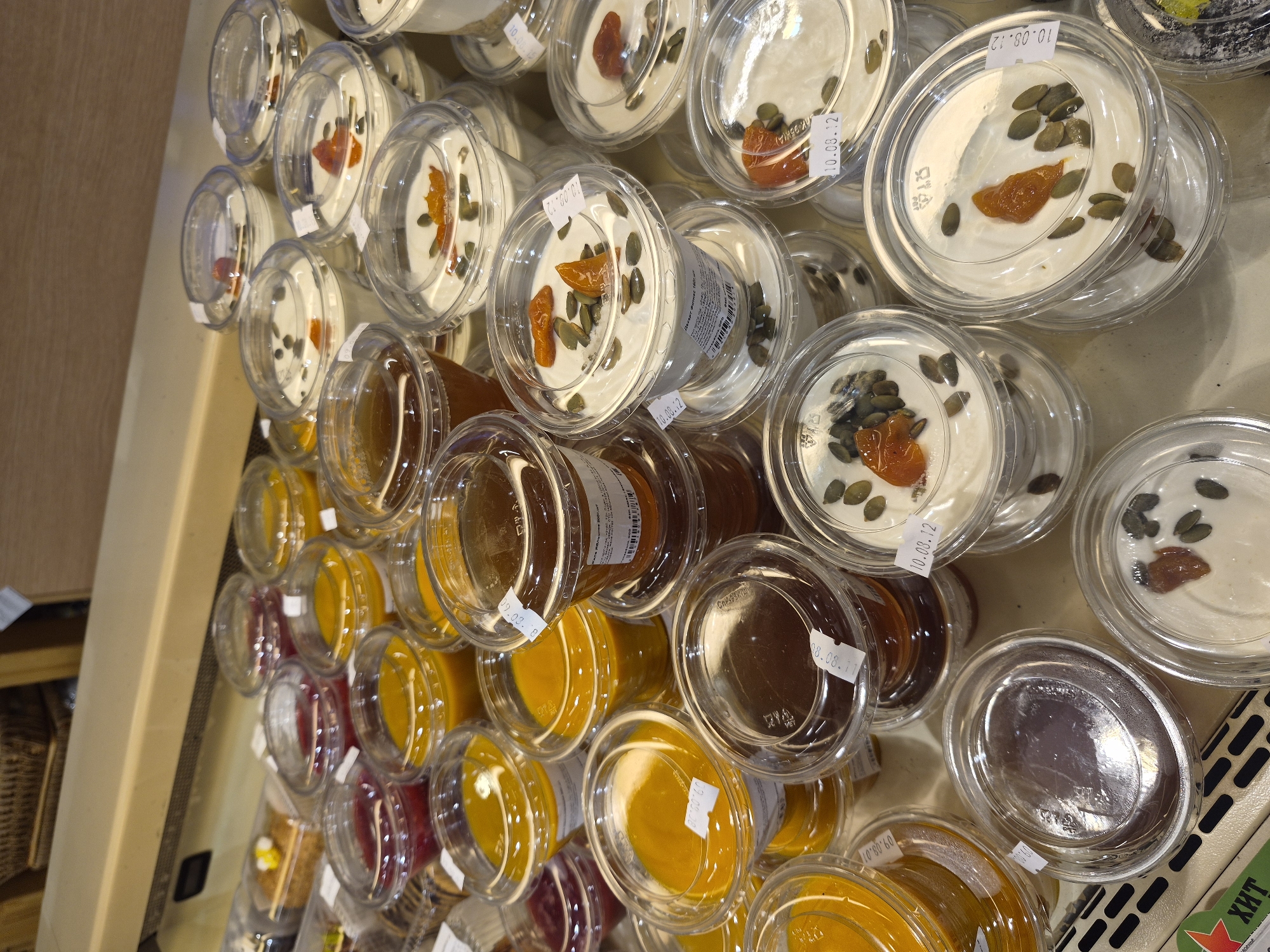 The image shows a display of individually packaged desserts and jams in clear plastic containers, arranged on a shelf in what appears to be a retail store or bakery in Yekaterinburg, Russia. The desserts are primarily creamy white with visible toppings of pumpkin seeds and dried fruit, possibly apricots. Several containers hold amber-colored liquids, likely jams or honey, and some contain a dark purplish substance. The containers are stacked and varied in size, with some featuring white labels with text, including dates such as "10.08.12". In the background, a woven basket and wooden shelving are partially visible. A green label with white Cyrillic text, "ХИТ", is visible at the bottom left. There are no people or animals in the frame. The lighting suggests an indoor, commercial setting.