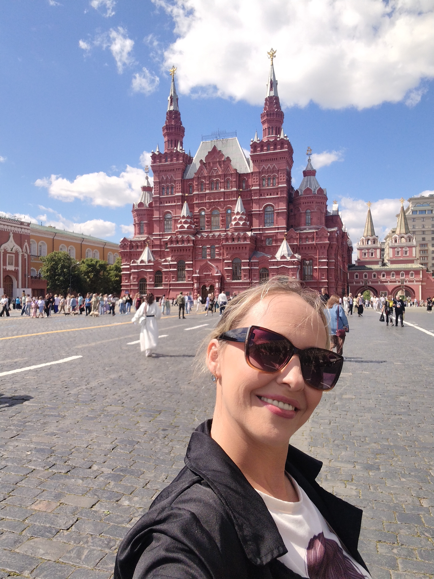 In the foreground, a woman with blonde hair and sunglasses smiles at the camera, her face filling the bottom right of the frame. She is wearing a black jacket over a white t-shirt with a graphic design. In the background, a large, ornate red building with intricate spires dominates the scene under a bright blue sky with scattered white clouds. People are visible walking in the plaza in front of the building, and more buildings and trees are visible in the distance. The ground is paved with cobblestones.