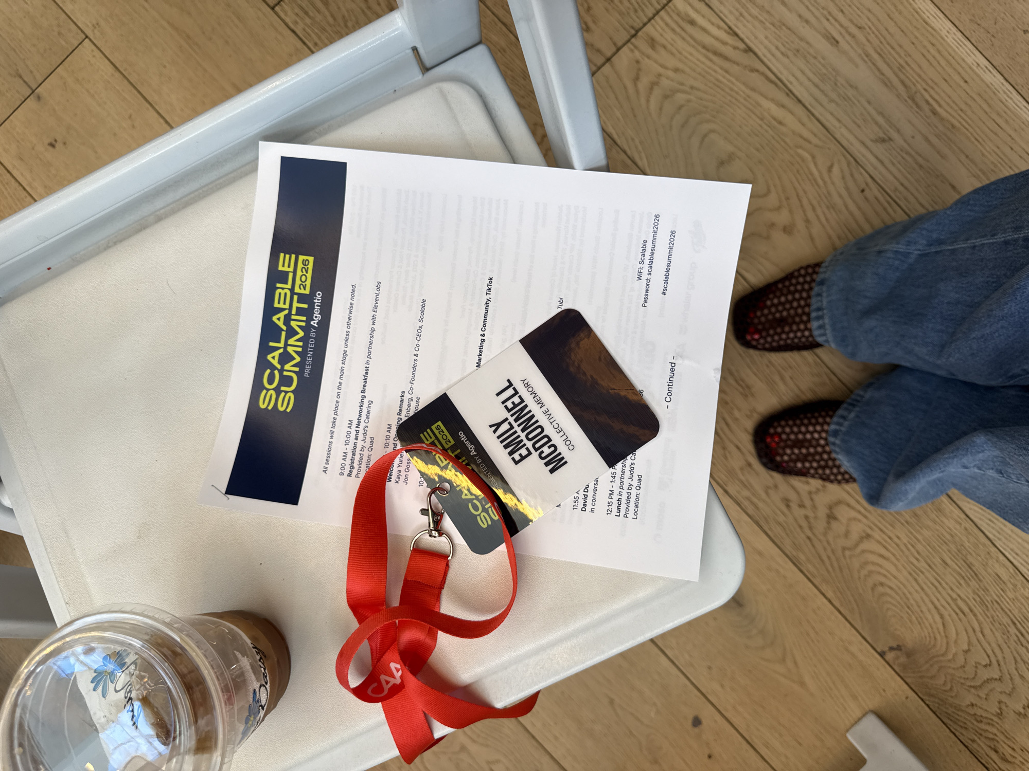 An overhead view captures an attendee's items resting on a white chair, likely during a professional event or conference. A red lanyard, featuring the "CAA" logo, is attached to a name tag for "EMILY MCDONNELL" from "COLLECTIVE MEMORY."
Beneath the lanyard and name tag lies a printed agenda for the "SCALABLE SUMMIT 2026 PRESENTED BY Agentio." The agenda outlines various sessions and activities, including "Registration and Networking Breakfast in partnership with ElevenLabs," "Welcol And Opening Remarks" by Kaya Yurien and Jon Goss, and "Lunch in partnership with Judd's Catering." Other visible details on the agenda include mentions of "Marketing & Community, TikTok," "Tubi," and speaker "David Du." Crucially, it provides "WiFi: Scalable" with the password "scalablesummit2026" and the hashtag "#scalablesummit2026."
To the left, a clear plastic cup containing an iced beverage sits on the chair. In the bottom right corner of the frame, a person's feet, clad in blue jeans and brown mesh-patterned shoes, are visible on a light wooden floor. The overall scene suggests a break or a moment of rest for an attendee at a multi-day summit.