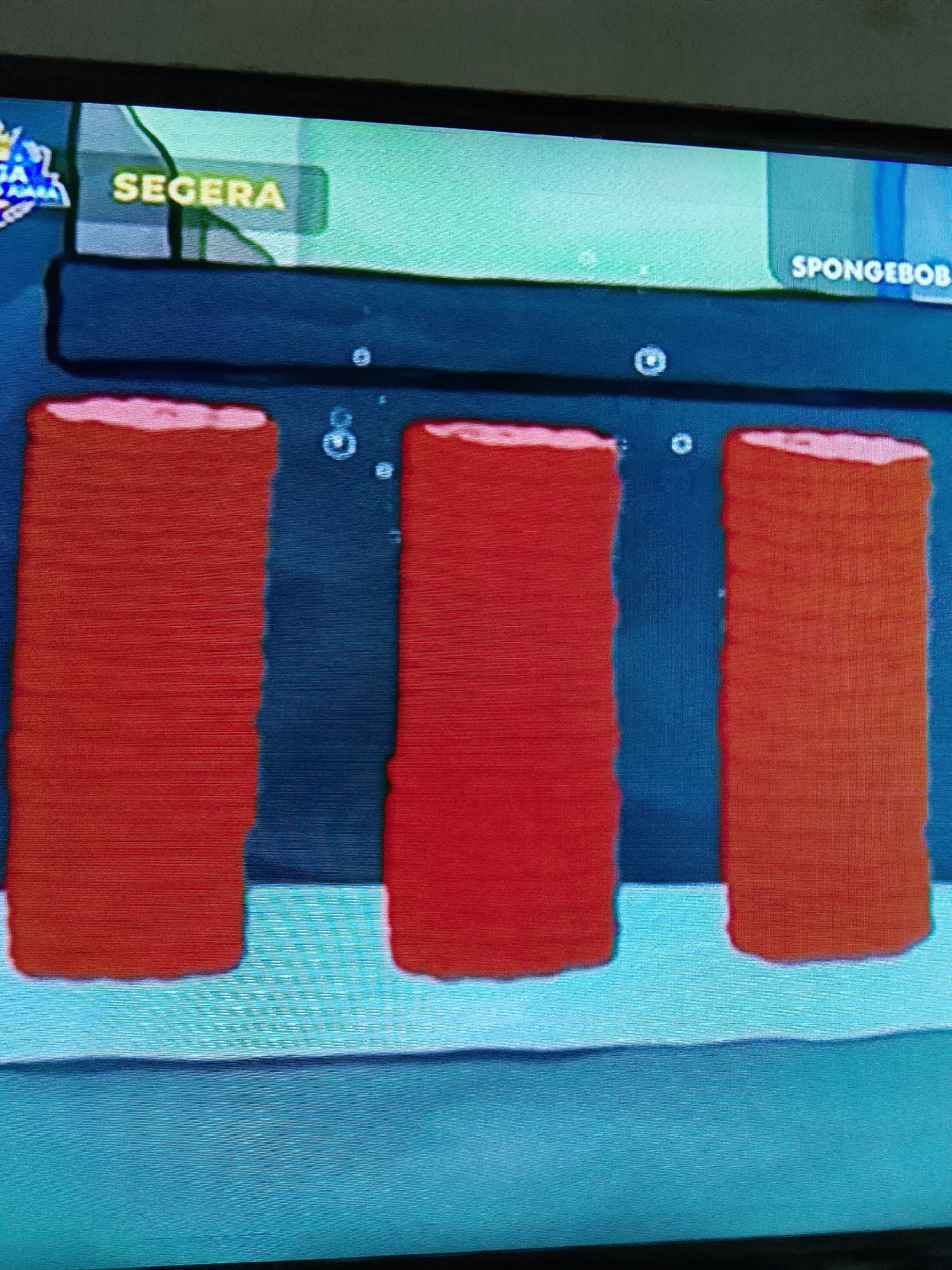The image displays a screenshot from a video game, likely a mobile or console game, featuring a stylized underwater environment. Dominating the foreground are three large, vertical, red cylindrical objects with pink tops. These objects appear to be arranged side-by-side with some spacing between them. The background features a dark blue textured area with scattered small circular bubbles, suggesting an aquatic setting. Above this, a lighter blue and green gradient forms the upper background. Two text elements are visible: "SEGERA" in a yellow rectangular box on the left, and "SPONGEBOB" in white text on the right, indicating the game's context. There are no people or discernible actions taking place. The image's origin in Merbopuntong, Indonesia, is not visually represented and cannot be determined from the provided content.