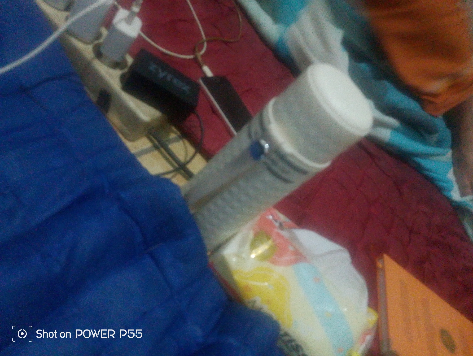 The image captures a cluttered indoor scene, likely a bedroom or living area, in Lebaksiu, Indonesia. A blue fabric drapes across the left side of the frame, partially obscuring a power strip with several electrical plugs and a black device labeled "Zyrex." A white cylindrical object, possibly a portable speaker or battery pack, is prominently positioned in the center, connected to a charging cable. A smartphone is also visible, plugged into the power strip. In the background, a maroon fabric covers a surface, with parts of a blue and white striped blanket visible. A person's orange-colored clothing is visible at the top right of the frame. The overall impression is one of everyday domestic activity, with devices being charged and various items casually arranged.