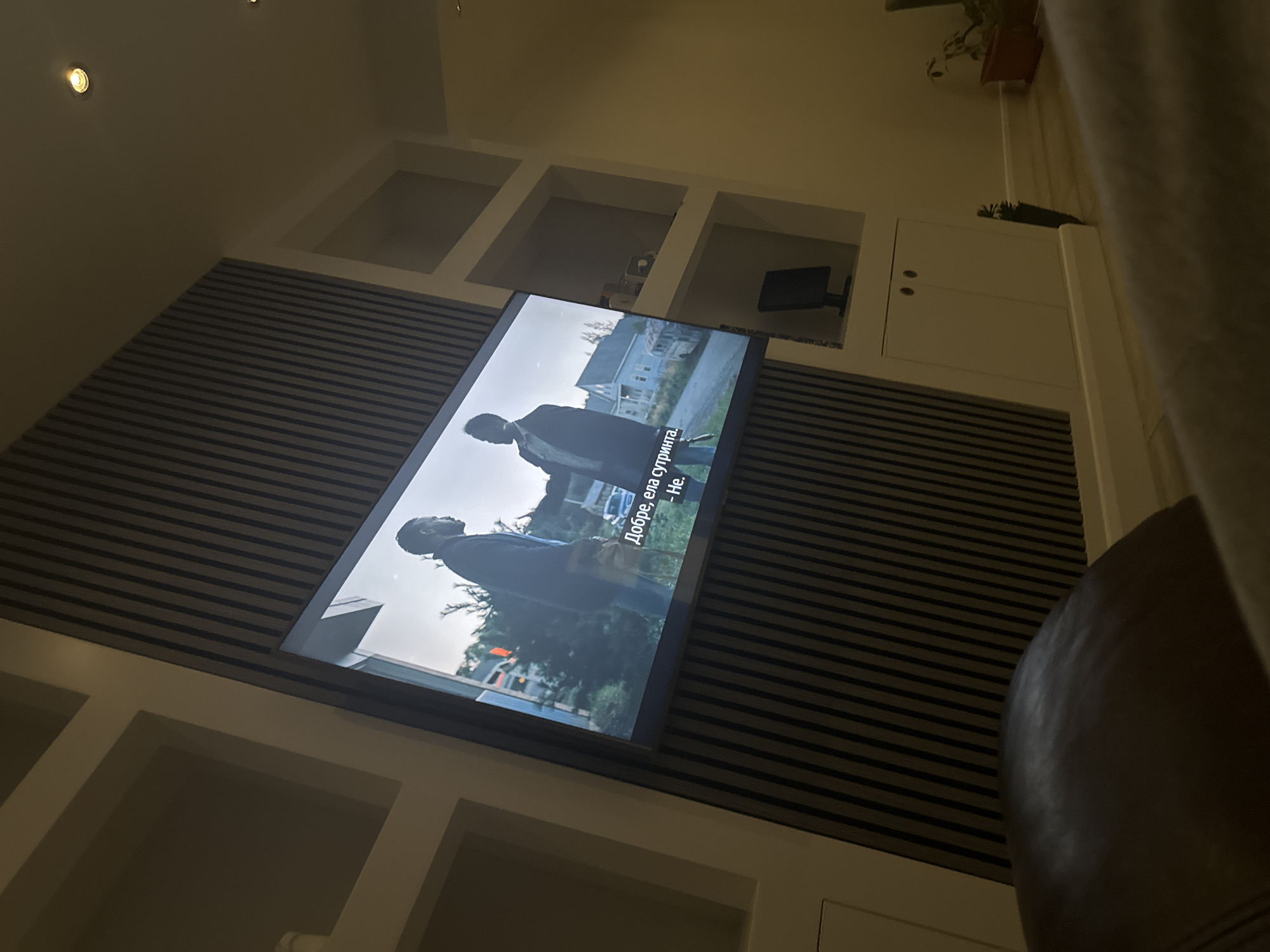 The image shows a television mounted on a wall with horizontal striped paneling. The TV screen displays a scene with two figures in a green, outdoor setting. Subtitles in a language other than English are visible. The wall features white, built-in shelving and cabinets. A chair is partially visible in the foreground. The image does not provide enough information to determine the location in the city of Chatham, United Kingdom.