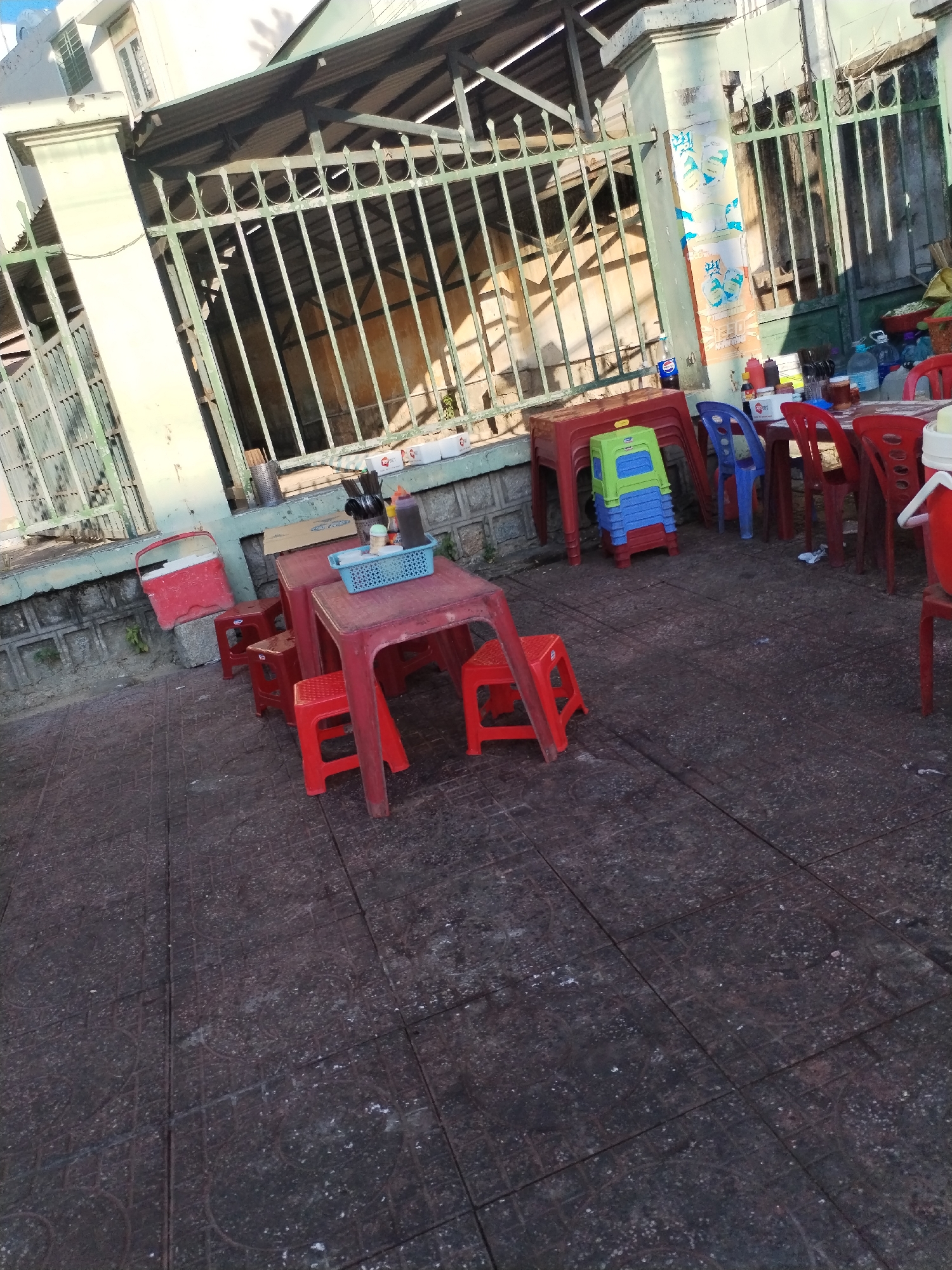 The image shows an outdoor seating area of a street food vendor in Bac Nha Trang Ward, Vietnam. The seating area consists of several red plastic tables and stools. There is also a stack of blue and green plastic stools. Table accessories include condiments and utensil containers. Behind the seating is a building and a metal fence. The street surface is paved with dark grey stone tiles.