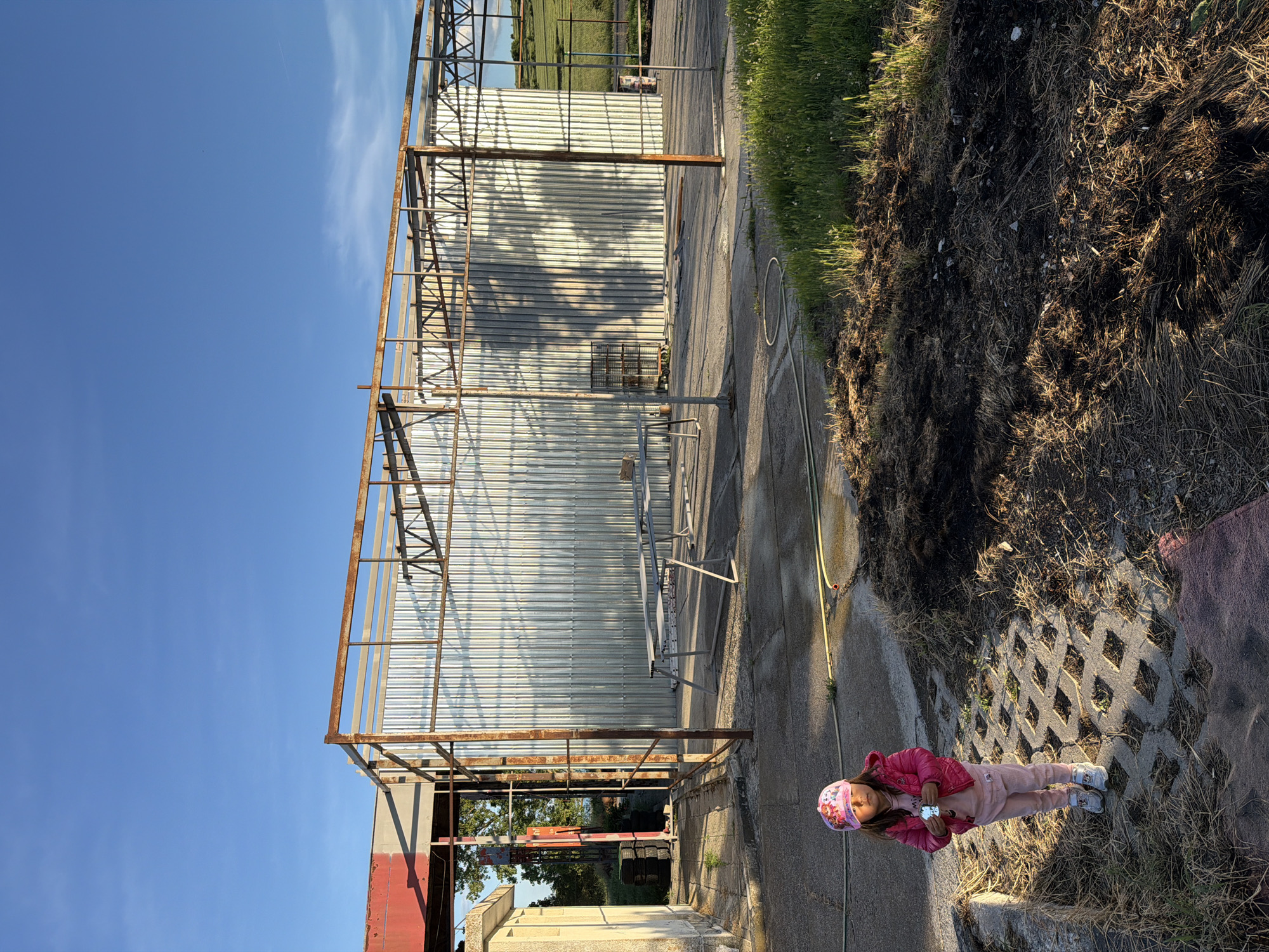 A low-angle outdoor shot in Dabnik, Bulgaria, shows a corrugated metal structure with a rusty metal frame against a blue sky. A young girl wearing a pink jacket, pink pants, and a pink hat stands on interlocking concrete blocks with grass and dry vegetation in the gaps. A yellow hose lies on the concrete walkway, leading toward the metal structure. Beyond the structure, a green field is visible. The foreground consists of dark soil and vegetation. The lighting suggests late afternoon or early morning.