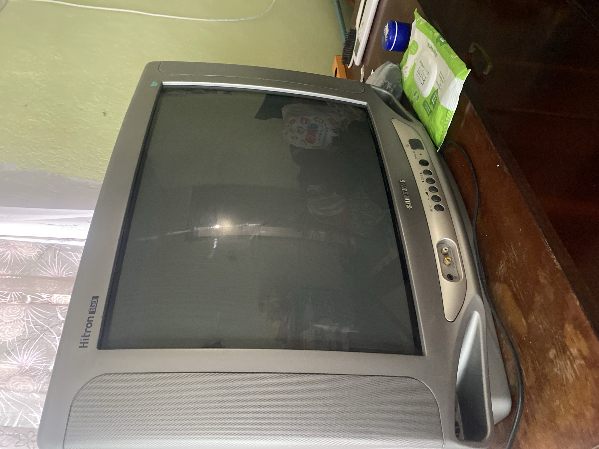 An older television, a "Hitron Black" model made by Samsung, sits on a wooden surface in a room with light green walls. The TV screen is dark, but reflects the surrounding environment. Several items, including a blue container and a package of baby wipes, are visible on the same surface as the TV. The location is Katuntsi, Bulgaria. No people are visible.