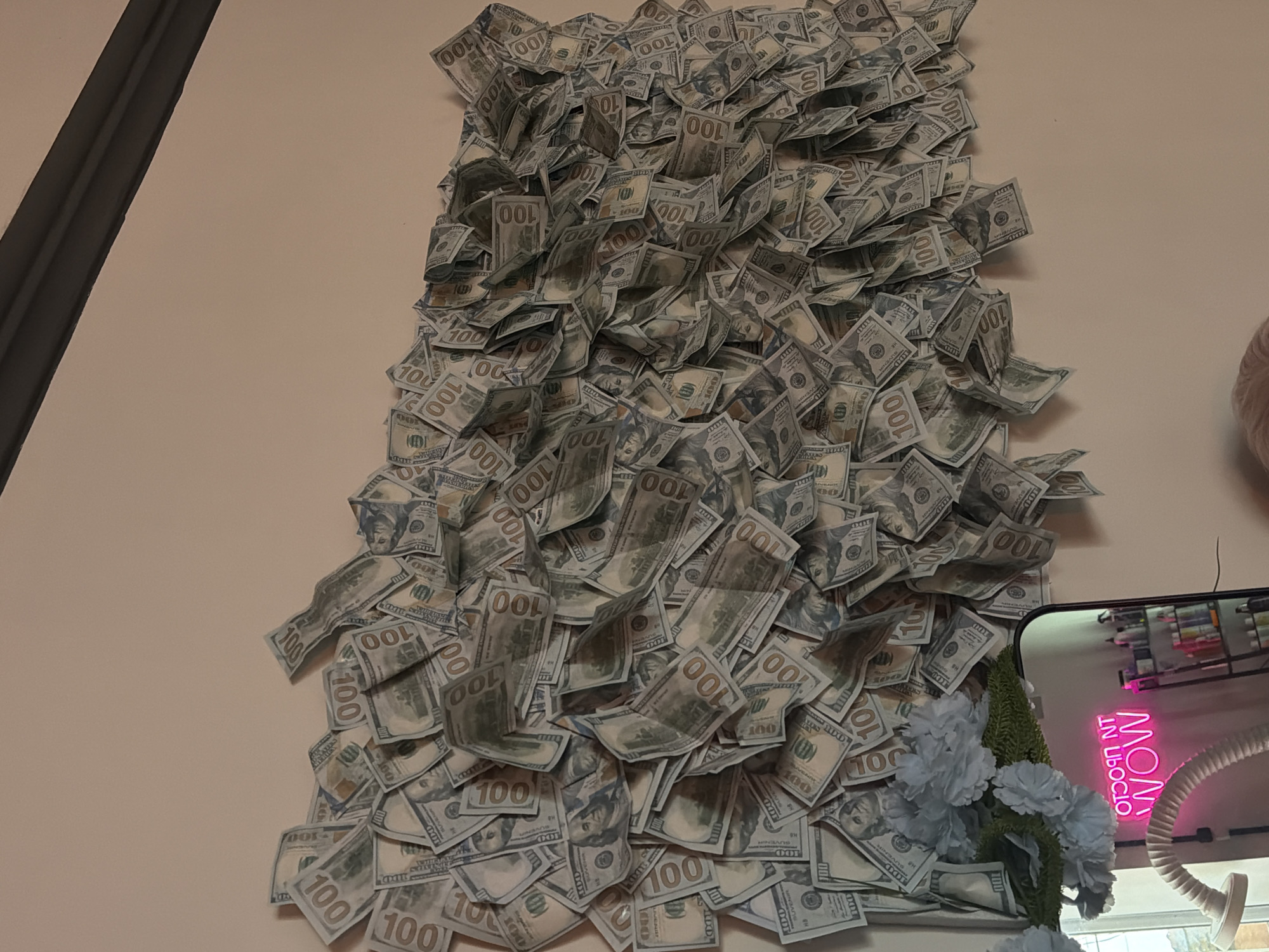 A wall is covered in a large display of US $100 bills. The bills are layered on top of each other, with some folded and others spread flat. In the lower left corner, a vase of light blue flowers and some greenery is visible, as well as a mirror reflecting a neon sign that reads "NOT WOW" in pink lettering. The mirror also shows a shelf with various bottles. To the right of the mirror is a dark shelving unit with more bottles. A white, flexible hose is also visible in the lower left of the image. The wall is painted a light, neutral color.