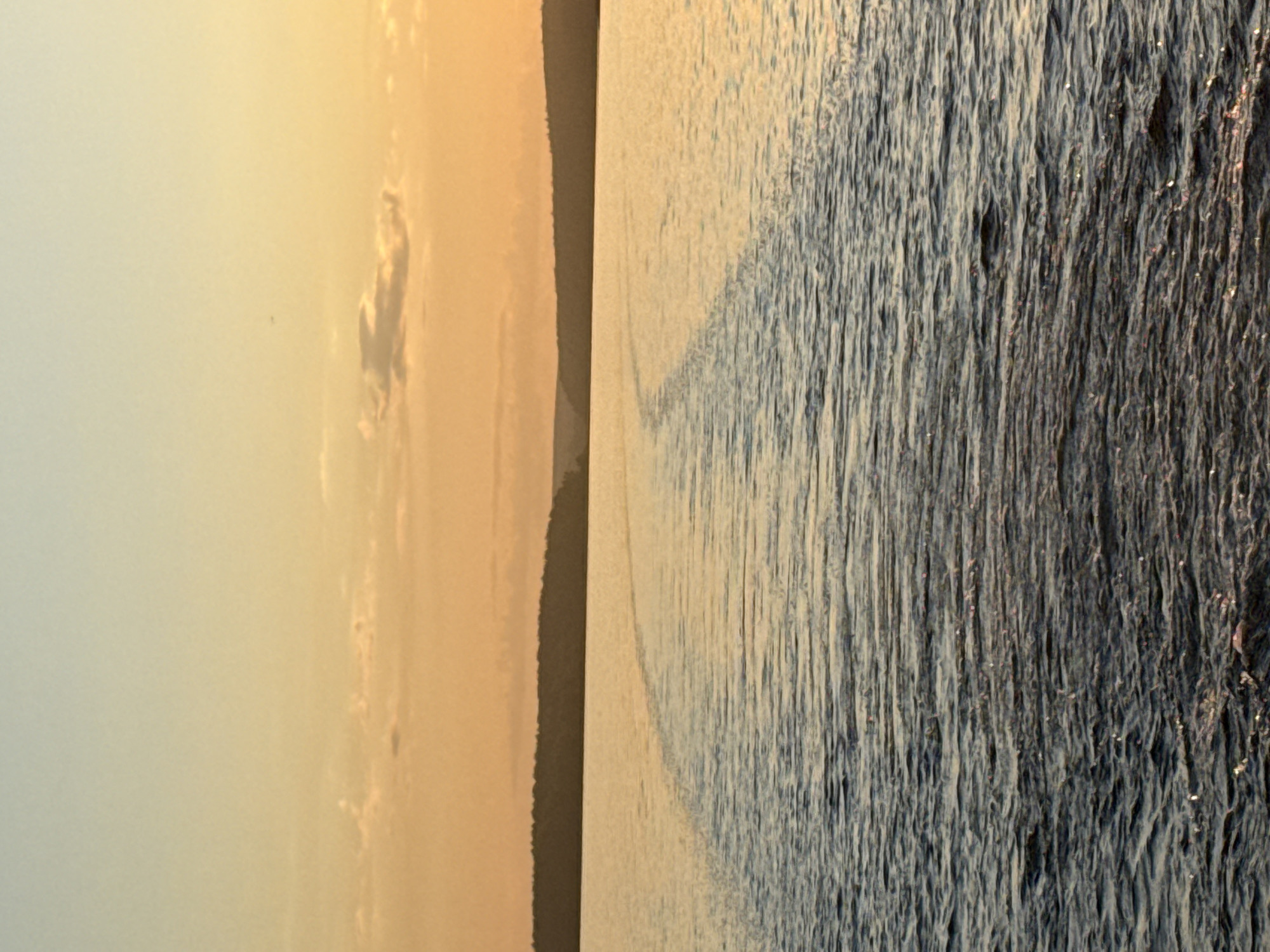The image depicts an outdoor waterscape at either sunrise or sunset, characterized by warm light. The upper portion of the frame features a sky transitioning from a pale blue-grey overhead to rich orange and yellow hues at the horizon. Sparse, diffuse clouds are present in the lower sky. A dark, continuous silhouette of a forested landmass, likely distant hills or a shore, spans the horizon line against the bright sky. The middle ground and foreground are dominated by a body of water, consistent with a large lake or reservoir. A prominent, curved wake or trail is visible on the water's surface, extending from the lower-left towards the upper-center, indicating the recent passage of a watercraft. This wake appears brighter than the surrounding water due to reflected light. The water in the foreground displays numerous small ripples and waves, with sun glints creating sparkling highlights on the crests. No individuals or explicit man-made objects, aside from the implied vessel, are present. The scene is consistent with a view of a large body of water near Votkinsk, Russia, such as the Votkinsk Reservoir.