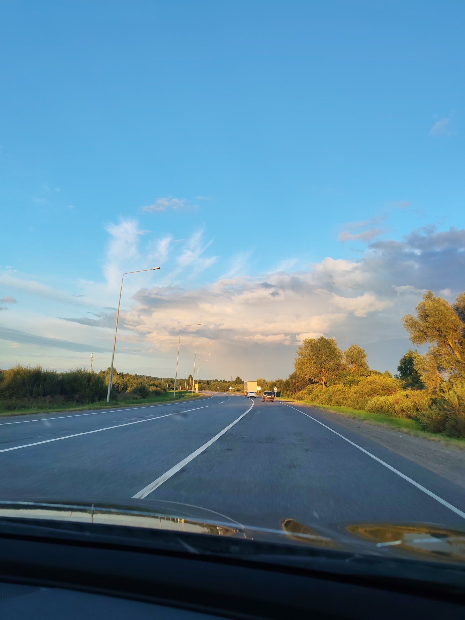 This is a photograph taken from the perspective of a vehicle moving on a highway. The road is asphalt with white lane markings. On either side of the road, there is lush green vegetation and trees. The sky is a vibrant blue with scattered white and gray clouds, some of which are illuminated by the setting sun. There are streetlights visible along the side of the road. In the distance, there are a few vehicles on the highway, including a truck and a car. The overall scene suggests a rural or suburban setting during golden hour. The location is the highway leading into the city of Gornyak, Russia.