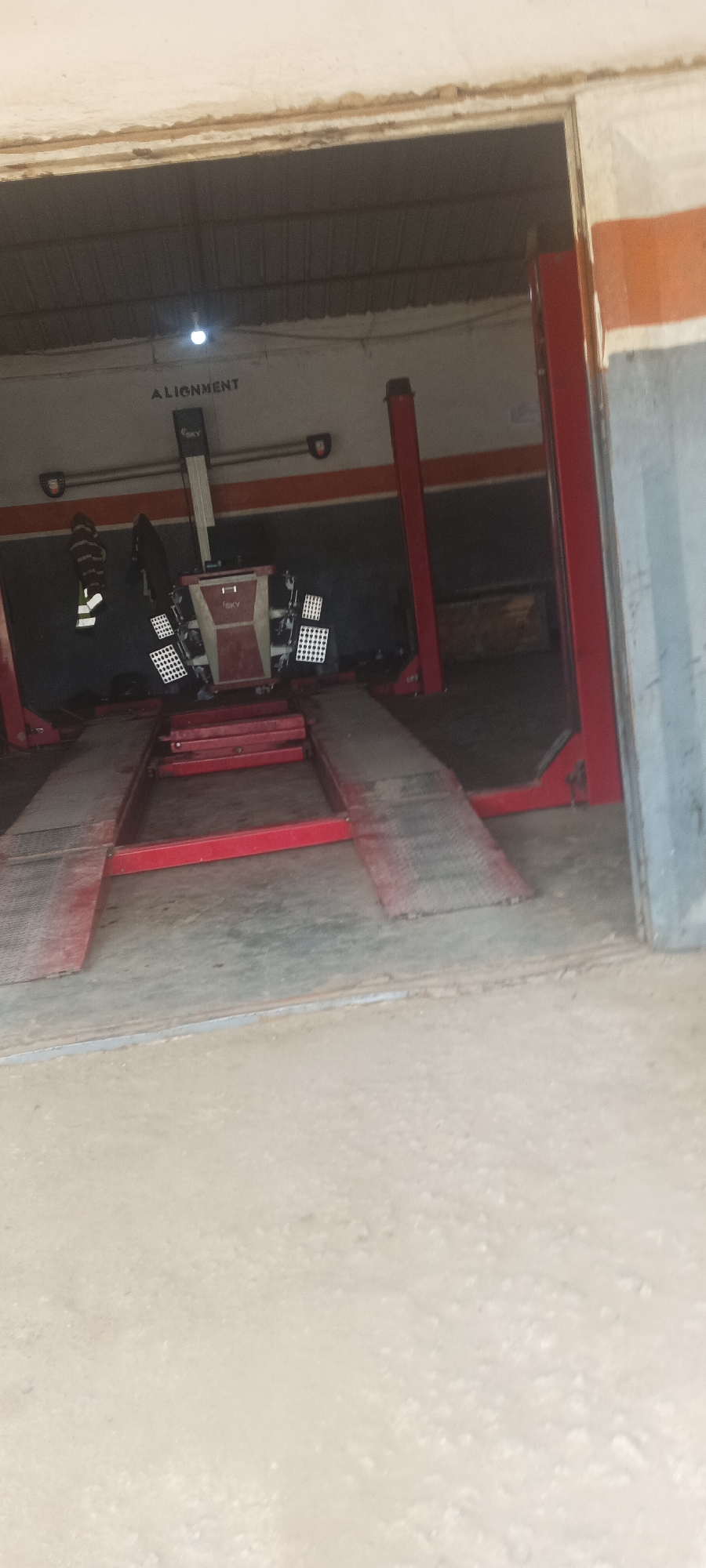 This image captures a car alignment bay within a workshop in Bukuru, Nigeria. The scene features a red hydraulic car lift with its platforms extended. Positioned on the lift is a car alignment machine, a complex device with multiple camera sensors and reflective targets attached to a central unit. The machine is branded with "iSKY". The wall behind the alignment machine is dark grey with a horizontal orange stripe. Above the wall, the word "ALIGNMENT" is stenciled in black capital letters. To the left of the alignment machine, there is a reflective vest hanging on the wall. The floor of the bay is concrete. The entrance to the bay appears to be open, with a dimly lit corrugated metal roof visible overhead. The overall impression is a functional space dedicated to automotive maintenance.