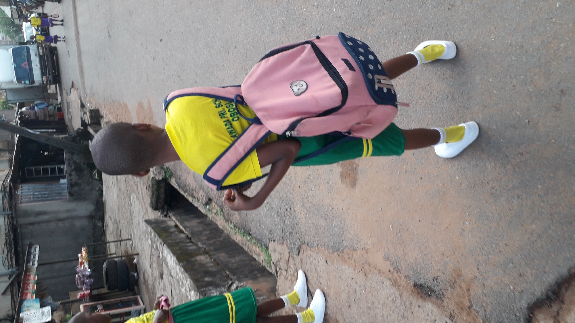 A young boy is walking on a street in Onitsha, Nigeria, with a pink backpack. He is wearing a yellow shirt, green shorts, white shoes, and yellow socks, apparently a school uniform. A second child is seen only from the knees down, wearing similar attire. There is a building and a drainage ditch to the side, and a white van is in the background. The environment appears to be a residential or commercial area.