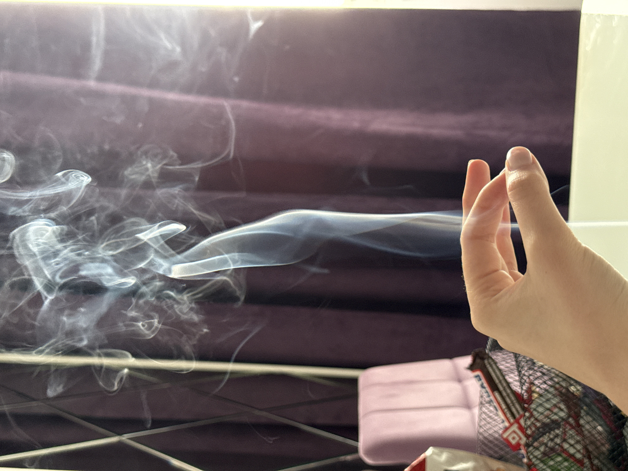 The image presents a close-up, indoor scene. A hand is positioned on the right side of the frame, with the thumb and index finger close together. Wisps of smoke emanate from the left side, drifting horizontally across the frame towards the hand. The background consists of a dark purple, possibly velvet-like material, with faint horizontal lines suggesting a padded headboard or similar structure. Below the smoke, there is a glimpse of a metal grid, a pink object (possibly a cushion), and packaging of unknown content.
The location is Unknown, Unknown.