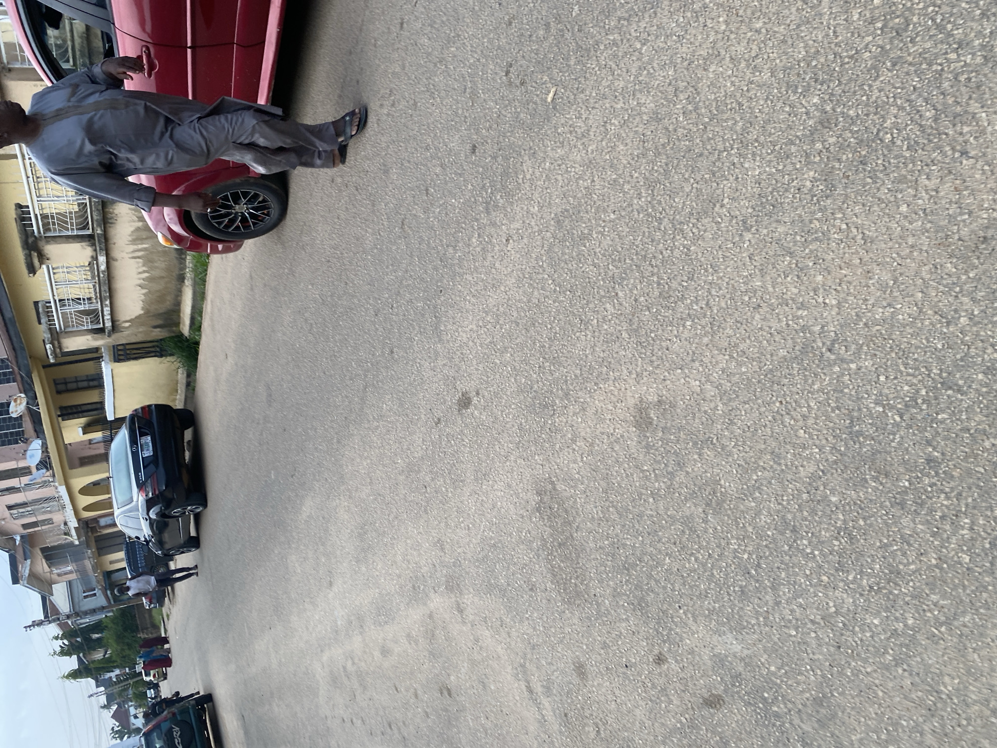 The image depicts a street scene in Jos, Nigeria. A person in a grey traditional outfit with sandals is walking on the asphalt road next to a red car. In the background, a black Mercedes-Benz sedan is parked on the side of the road, and other vehicles and buildings are visible. The environment suggests a residential or commercial area within the city. The overall impression is of a typical day in a Nigerian urban setting.