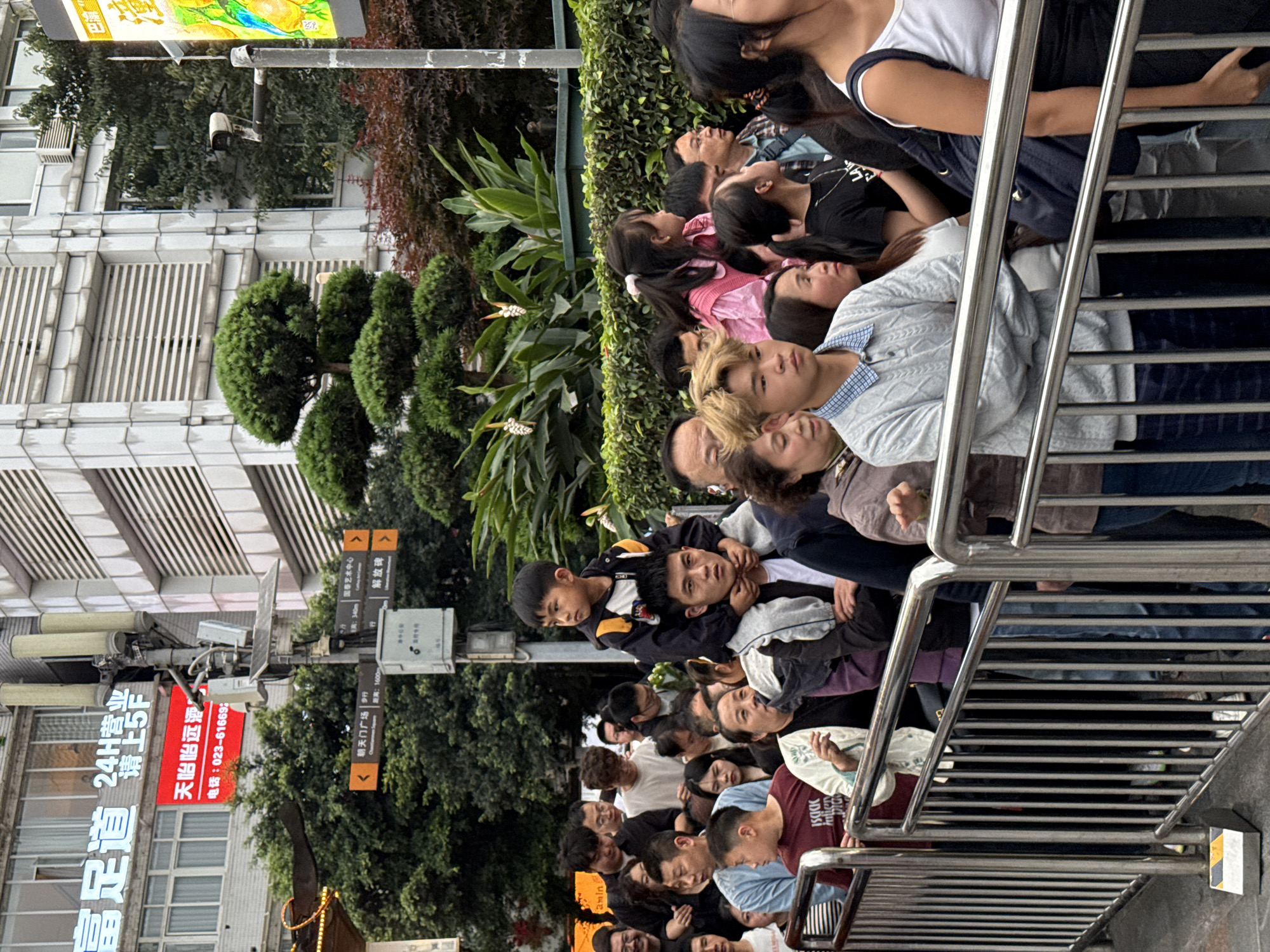 A medium shot captures a large crowd of people on an elevated pedestrian walkway in Yuzhong District, China. The walkway is lined with a metal railing, partially obscuring the people. The crowd, composed of individuals of various ages and ethnicities, is closely packed, with many looking upwards, presumably toward an unseen point of interest.
On the left side of the frame, buildings rise into the sky, including one with a prominent sign featuring Chinese characters and phone numbers. Street signs and greenery are interspersed between the structures, including what is identified as the Cathay Art Center. The general environment suggests a bustling urban setting.