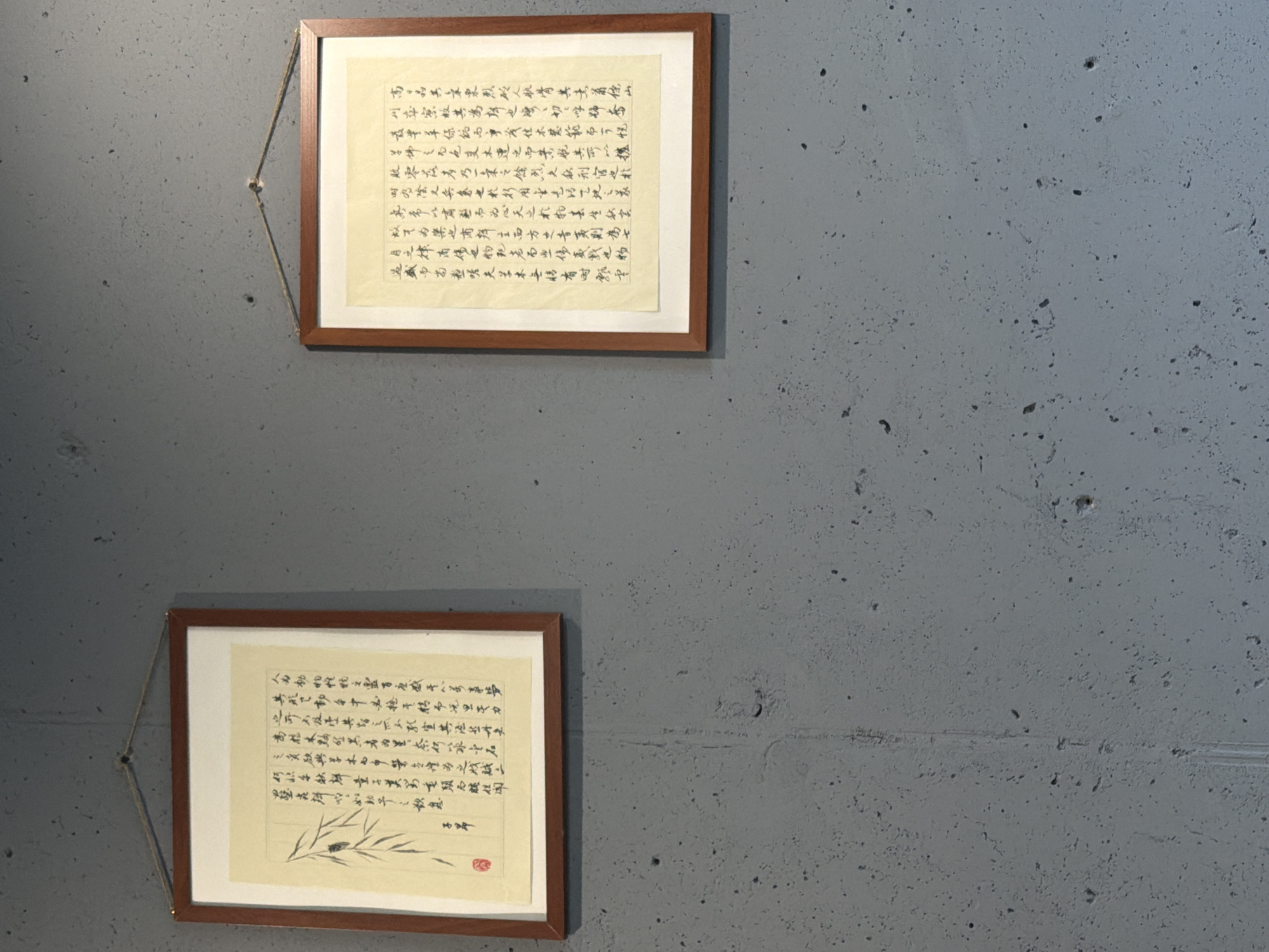 Two framed calligraphy pieces hang on a rough, gray concrete wall. The upper frame contains a dense text in traditional Chinese script, while the lower frame also features calligraphy, accompanied by a sparse drawing of bamboo leaves. The frames are dark wood, and the pieces are hung using cords. The overall environment is simple, suggesting a modern or minimalist setting. The location and city are not discernible.