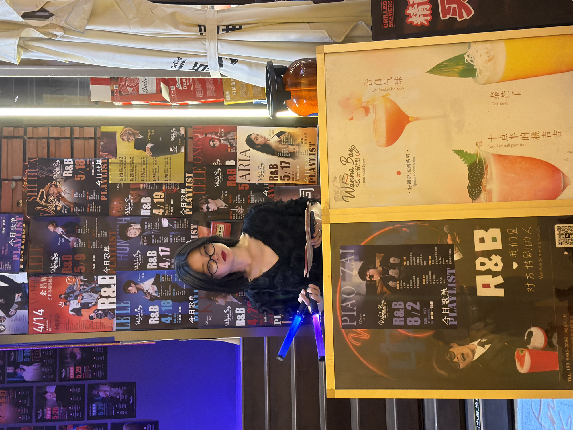 In a dimly lit establishment, possibly a bar or club, in Haizhu District, China, a woman with short black hair and glasses stands behind a yellow framed display. She is wearing a black, textured top and holds two blue light sticks in her hands, one glowing brighter than the other. She is also holding a folded piece of paper. Behind her, a wall is covered with numerous posters advertising music events, featuring artists and dates. The posters are predominantly in red, white, and black, with some featuring images of singers. In front of her, another yellow framed display features a cocktail menu with images of three drinks and accompanying Chinese text. To the right of the woman, a cream-colored umbrella with black stripes stands upright. Further to the right, a dark red sign with Chinese characters is visible. The overall environment suggests a place focused on live music and beverages.