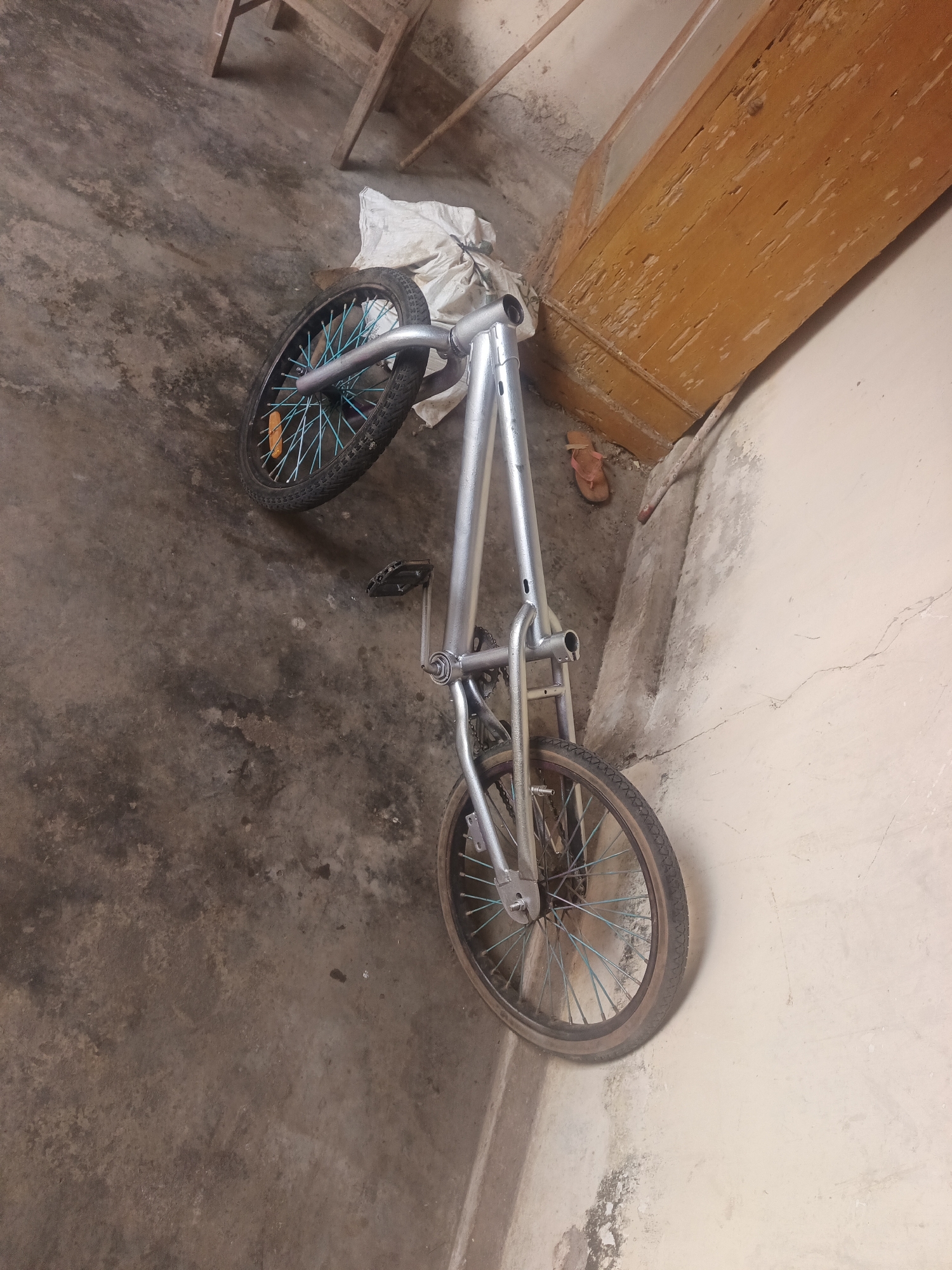 A silver bicycle, with blue spokes on both wheels, is leaning against a light-colored wall. The bicycle is positioned diagonally, with the front wheel higher than the rear wheel. A white cloth is draped over the handlebars. A single brown sandal is lying on the concrete floor near the front wheel. The floor is rough and stained. To the upper left of the bicycle, a wooden structure, possibly a chair or table, is partially visible. The wall behind the bicycle appears cracked and weathered. The scene is indoors, with overhead lighting casting shadows. The image was taken in Terbanggi Marga, Indonesia.