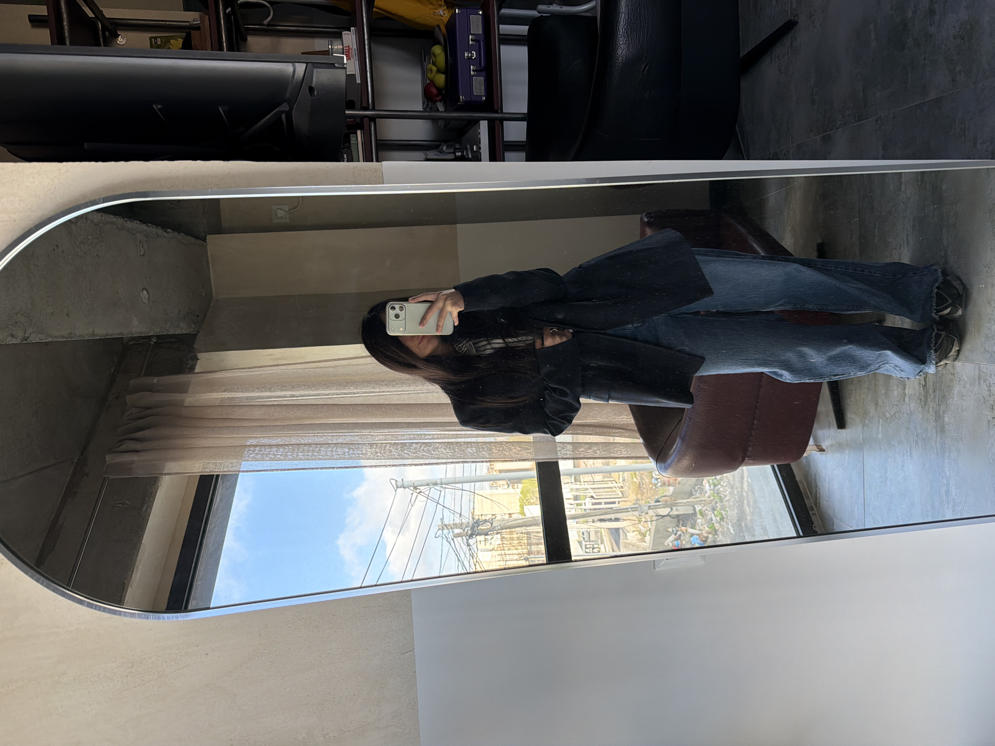 The image is a mirror selfie of a person standing in front of a mirror. The person is holding a smartphone and facing the mirror, capturing their reflection. They are wearing jeans and a black coat. The reflection shows a partial view of the room, including a window with a view of a cityscape with buildings and overhead cables. A brown chair is visible in the reflection, near the bottom-right of the image. The mirror is positioned near a shelf holding different items, and a portion of what appears to be a black chair is also in view.
Due to the view outside the window including buildings and power lines, it can be inferred that the location is in an urban area, most likely Yerevan, Armenia, based on the user's prompt.
