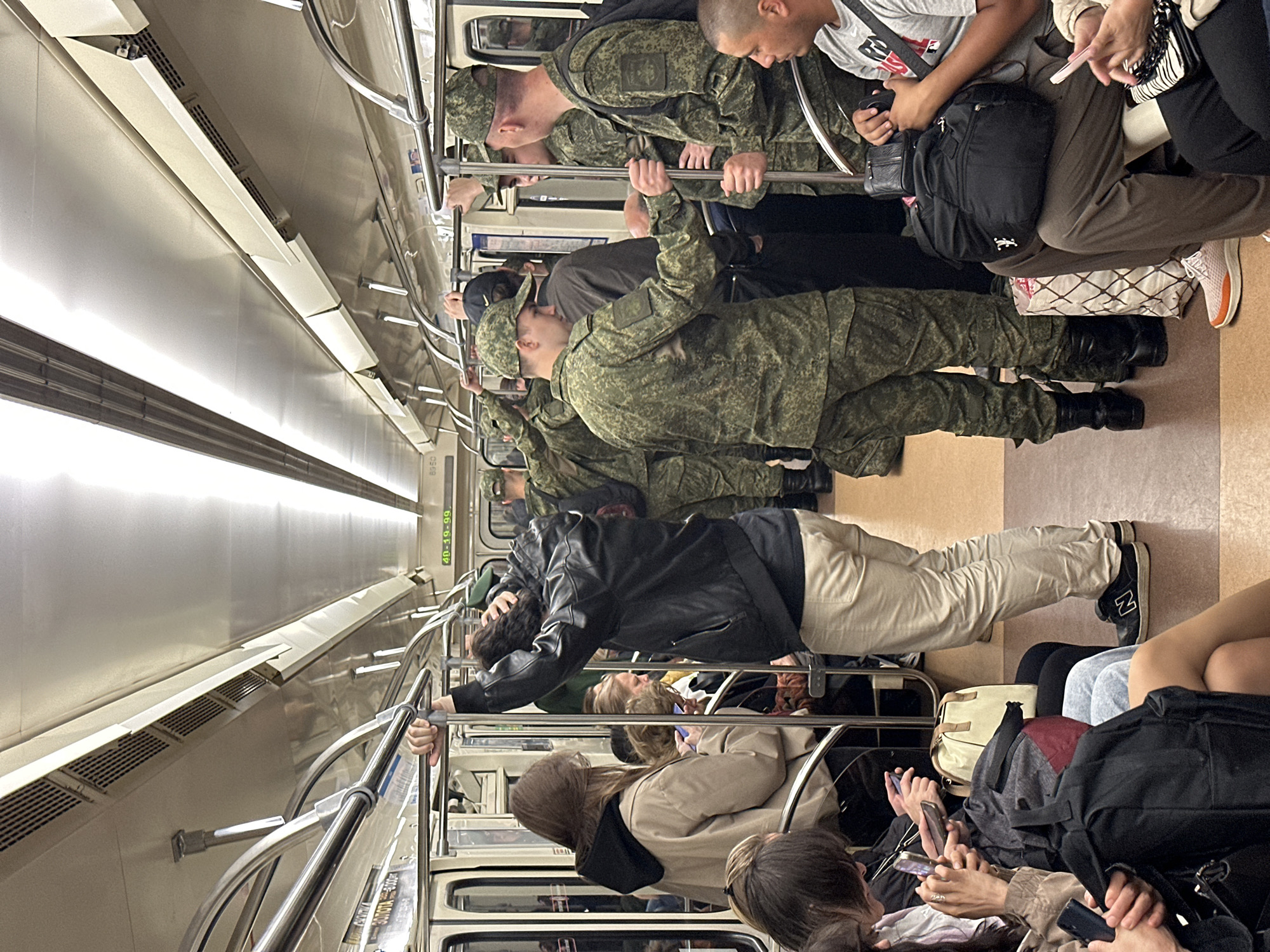 Inside a Saint Petersburg Metro train car, a group of people, including several individuals in military camouflage uniforms, stand and sit holding onto overhead rails and bars. The train car's interior is utilitarian with light-colored walls, ceiling, and lighting fixtures. On the right side of the car, a man in a grey t-shirt sits with a black backpack on his lap. In the center of the car, a man in a black leather jacket stands with his hand on a vertical pole, while a woman with long brown hair stands next to him, looking at her phone. Several other individuals, some in civilian clothing and others in military uniforms, are visible throughout the car. One soldier in camouflage is speaking to another soldier, who is holding onto a rail. Bags and backpacks are scattered on the floor and seats. The overall impression is of a crowded public transport setting.
