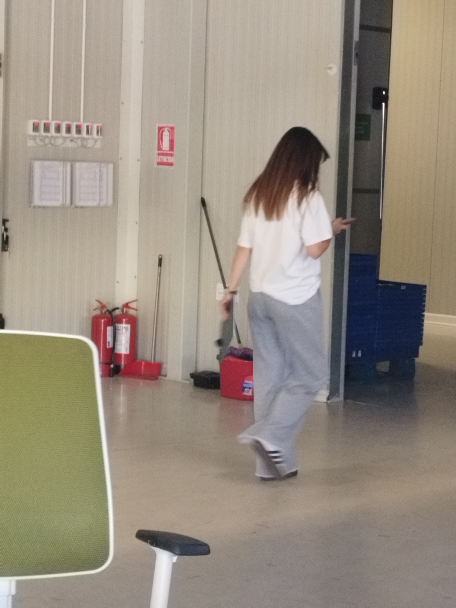 A woman with long, brown hair is walking away from the camera in a brightly lit, industrial-style room. She is wearing a white t-shirt and grey sweatpants, and white sneakers with black stripes. The room has light-colored, paneled walls. To the left of the woman, there are two red fire extinguishers and a red dustpan with a broom leaning against it. Above the fire extinguishers, there are two white boards mounted on the wall. Further to the left, there is a white panel with a row of small, square devices with red lights. To the right of the woman, there is a dark blue cart with multiple shelves. A green chair is partially visible in the foreground on the left. The floor is a light grey concrete. The overall environment appears to be a utility area or storage space.