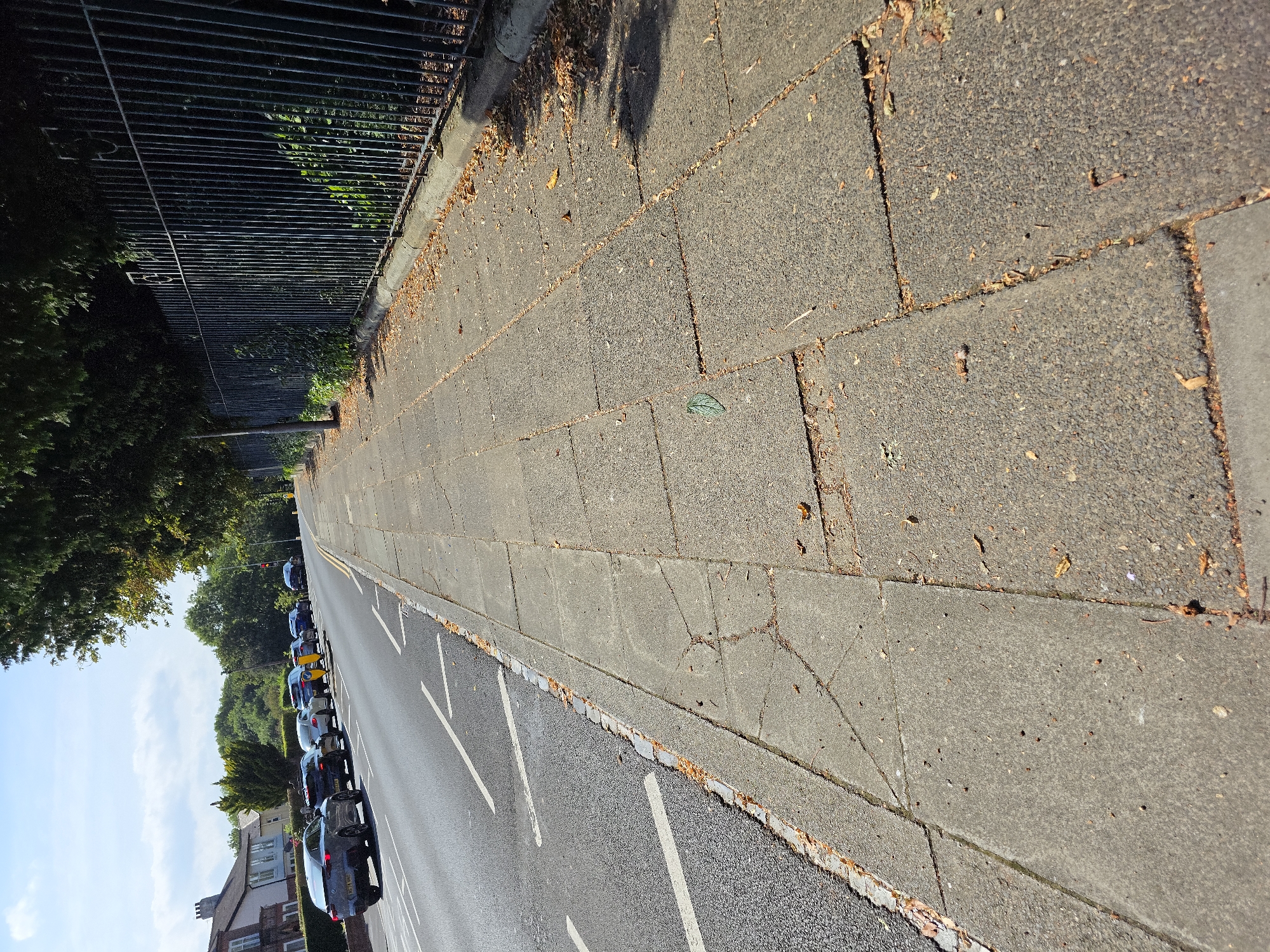 The image shows a view down a paved sidewalk in Stockton-on-Tees, United Kingdom, bordered by a road with several cars parked and driving, and a tall metal fence. The sidewalk is made of concrete paving slabs, some of which are cracked and have fallen leaves scattered on them. The road has white lane markings and a granite kerb separating it from the sidewalk. Beyond the fence, trees are visible, and the sky is blue with some clouds. The cars on the road appear to be parked in a residential or commercial area, given the houses visible on the left side of the road in the distance.