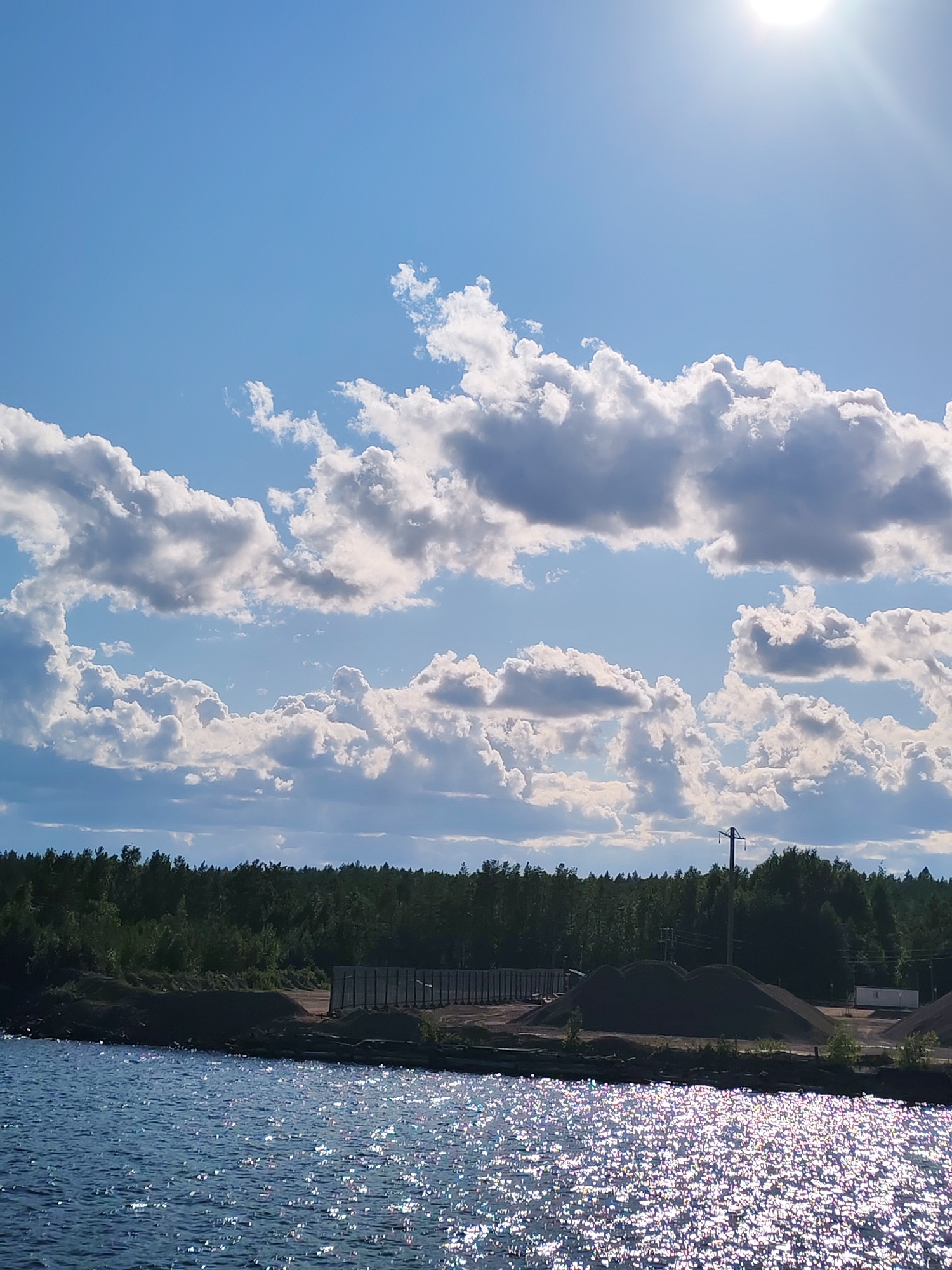 The image shows a body of water in the foreground with the sun glinting off its surface. In the midground, there is a treeline of dense, dark green forest. Behind the trees, on the shore, are piles of dark material, possibly gravel or sand, and some industrial structures including what appear to be shipping containers and a single utility pole with wires. The sky is a bright blue with scattered, fluffy white and gray clouds. The scene is likely a view of the industrial area near Kvartsitny, Russia, adjacent to a body of water. There are no people visible in the image.