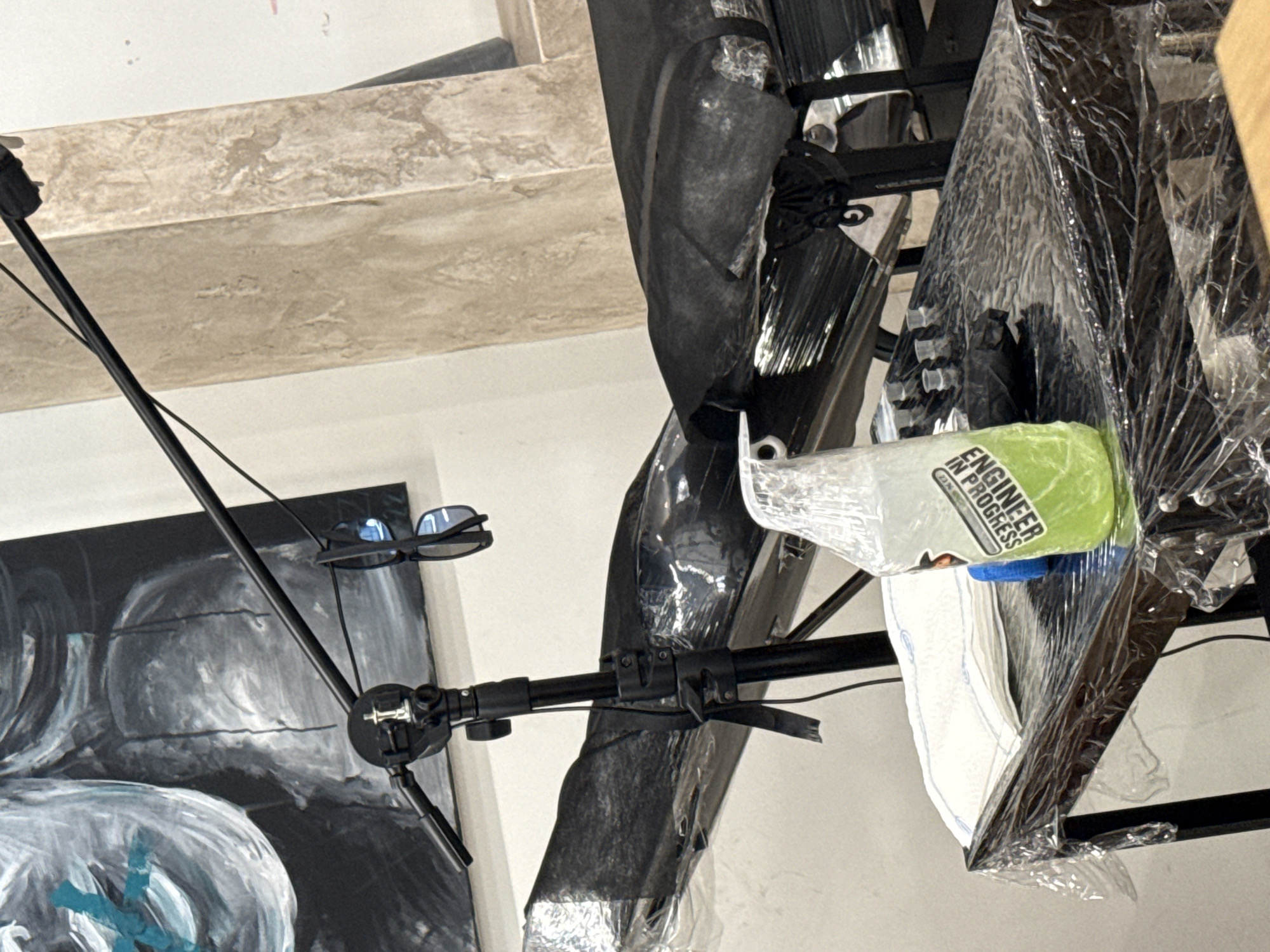 A medium-shot presents an indoor scene. An abstract art piece, primarily in black and white, with blue accents, is visible in the lower-left corner. A pair of sunglasses are perched on a black metal stand, which also holds an articulated black microphone arm. The stand's position suggests it is used for recording or broadcasting. Adjacent to the microphone, wrapped in plastic, is a framed artwork or photograph. A water bottle with a label that reads "ENGINEER IN PROGRESS" is also present. The environment has an industrial or studio feel, with a concrete ceiling and a general lack of domestic décor. The location cannot be determined from the objects in the image.
