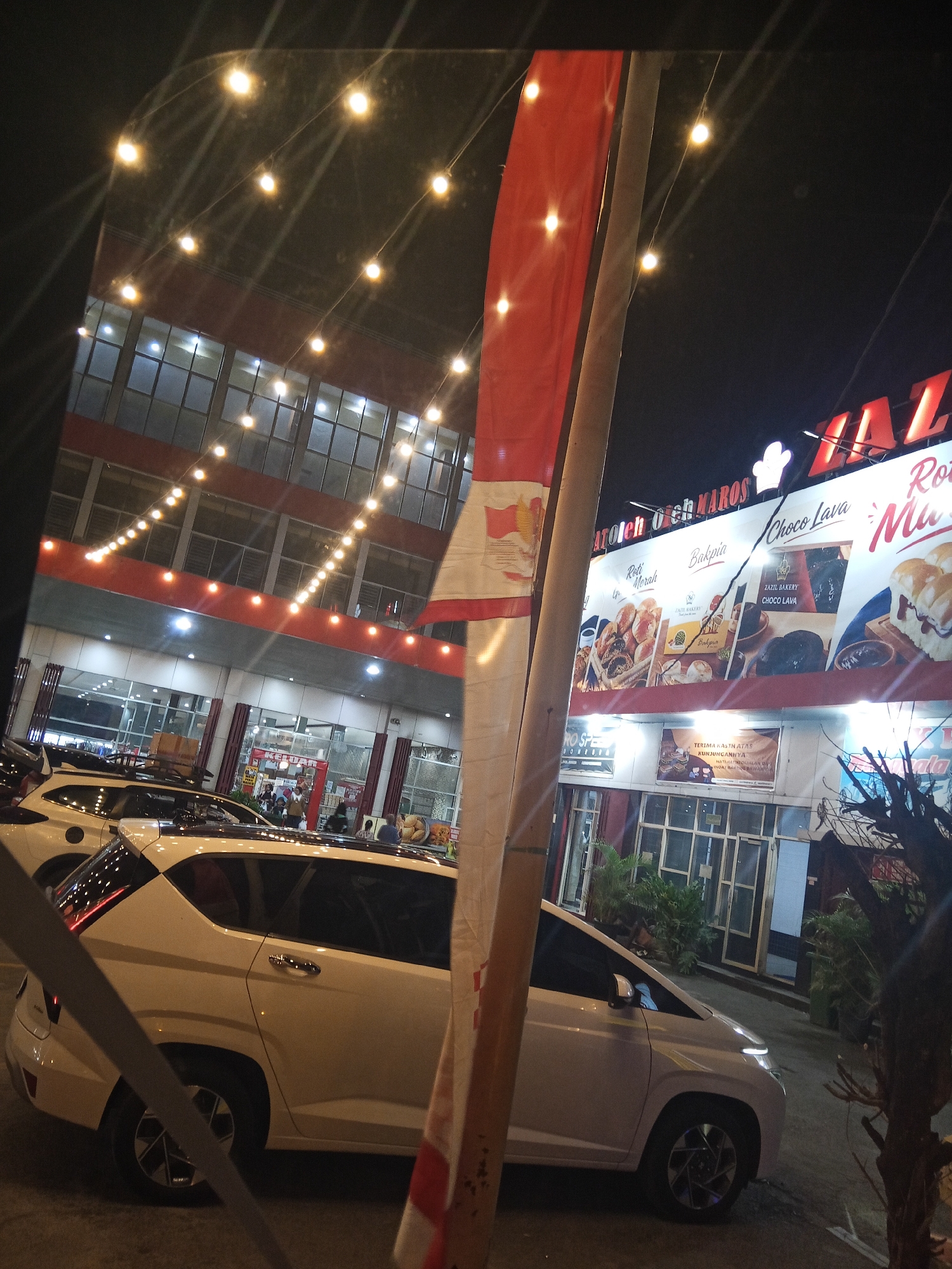 The image captures a street view at night in Hasanuddin, Indonesia, with a focus on a commercial building and parked vehicles. A white car is prominently positioned in the foreground, its rear on the left and its front on the right. Beyond the car, a multi-story building with illuminated windows stretches across the background. The building's facade features a large sign with red lettering that reads "HAZIL" above other text and images advertising baked goods, including "Choco Lava" and various types of bread. String lights are visible, illuminating the area and reflecting off the cars and building. Several people are visible near the entrance of the building, and other cars are parked nearby. A red and white flag or banner hangs from a pole on the right side of the image. The overall scene suggests a busy commercial area during the evening hours.