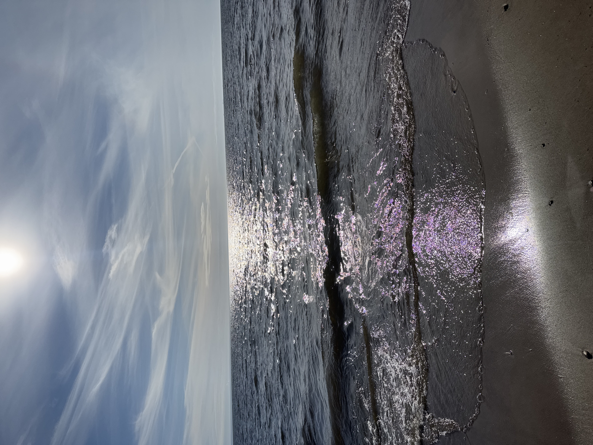 The image depicts a beach scene in Kronstadt, Russia, with the sun shining brightly in the upper left corner of the frame. The sky above is mostly clear with scattered wispy clouds. The sea occupies the center of the image, its surface rippled and reflecting the sunlight. The shoreline is visible on the right side of the image, with a sandy beach area. The sand appears wet and smooth near the waterline. There are no people visible in the image.
