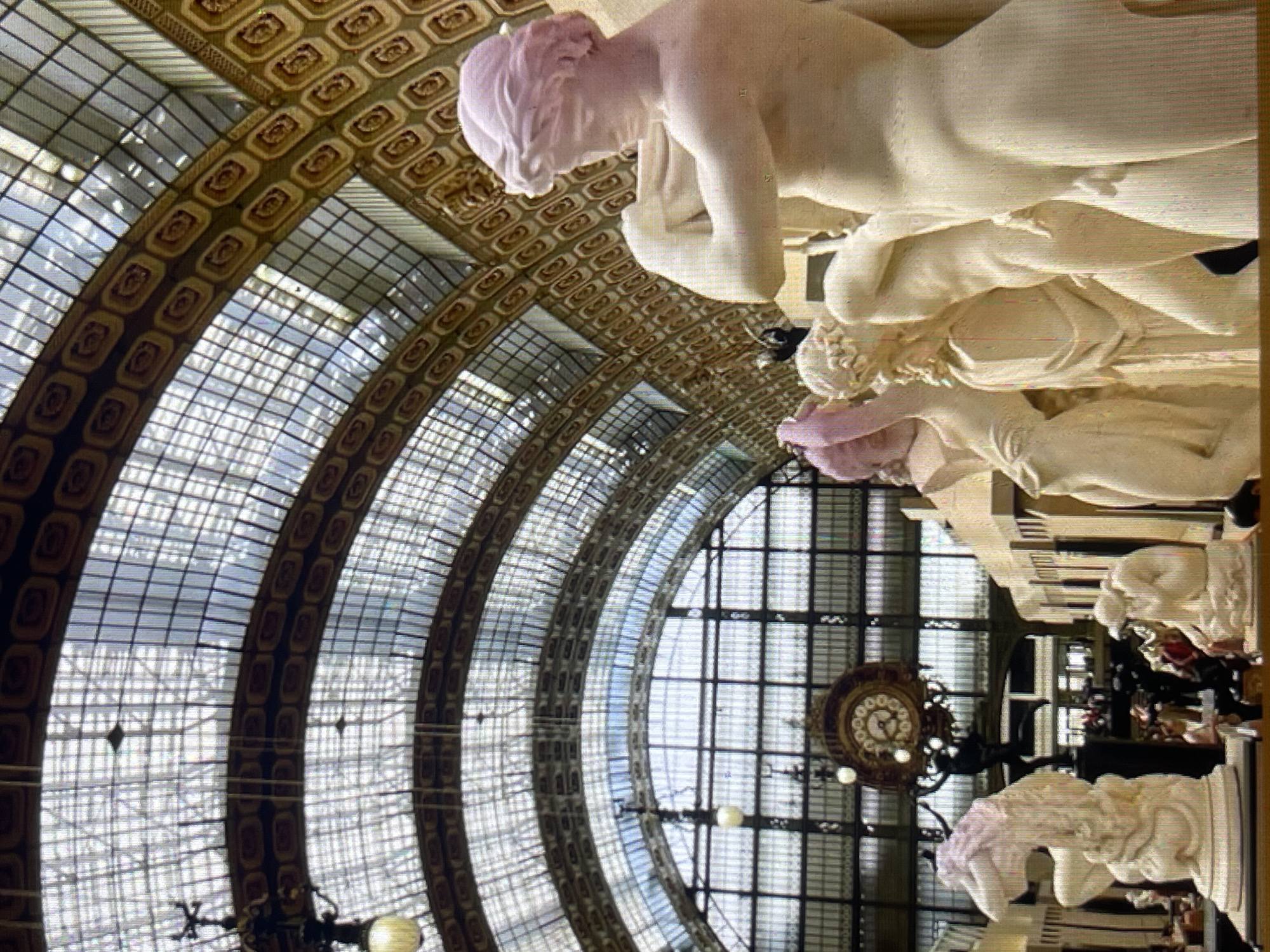 The image shows the interior of a grand hall with a glass and metal arched ceiling. In the foreground, there are several classical-style white marble statues, some with pink accents on their hair. In the background, a large circular clock hangs from the ceiling. People can be seen walking and observing the exhibits in the distance. The architecture suggests a museum or a large public building. The scene is well-lit by natural light filtering through the ceiling and artificial lights. There are no specific details in the image that would indicate the location in Bishkek City, Kyrgyzstan.